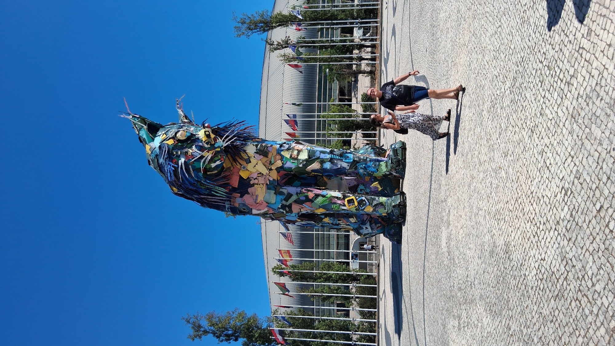 This is a full-shot, eye-level, outdoor, daytime photograph of a large, colorful sculpture of a lynx in front of a modern building with many flags. The sculpture, made of recycled materials, dominates the center of the frame. Its head is raised and tilted slightly to the right, with sharp, pointed ears and a fierce expression. Its body is a mosaic of brightly colored pieces, and its legs are thick and sturdy. Two people are walking on the paved ground in front of the sculpture. A woman on the left is wearing a long, patterned dress and a dark backpack. A man on the right is wearing a black t-shirt with a graphic and blue shorts. Behind the sculpture, a white building with a curved roof is visible, and a line of flags on poles stretches across the middle ground, separating the paved area from the building. The sky is clear and blue. The sculpture is located at the Parque das Nações, specifically in front of the Altice Arena, in Lisbon, Portugal.