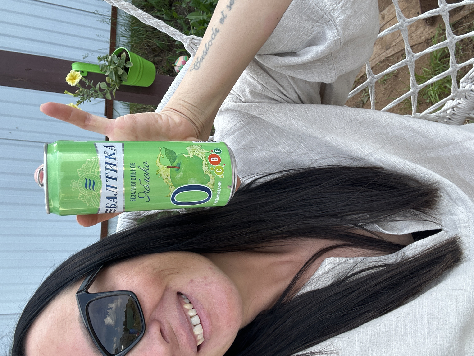 A medium close-up shot shows a woman outdoors, holding a can of "Baltika" non-alcoholic apple drink. She has dark hair, is wearing sunglasses with black frames, and a light-colored linen shirt. The can is green with white lettering and a picture of an apple. Behind her, a light blue fence and a brown wooden post with a small green planter holding yellow flowers can be seen. The woman is sitting in what appears to be a hammock, indicated by the white ropes and netting. Her left arm, visible in the right side of the frame, has a tattoo. The background suggests a backyard or garden setting.