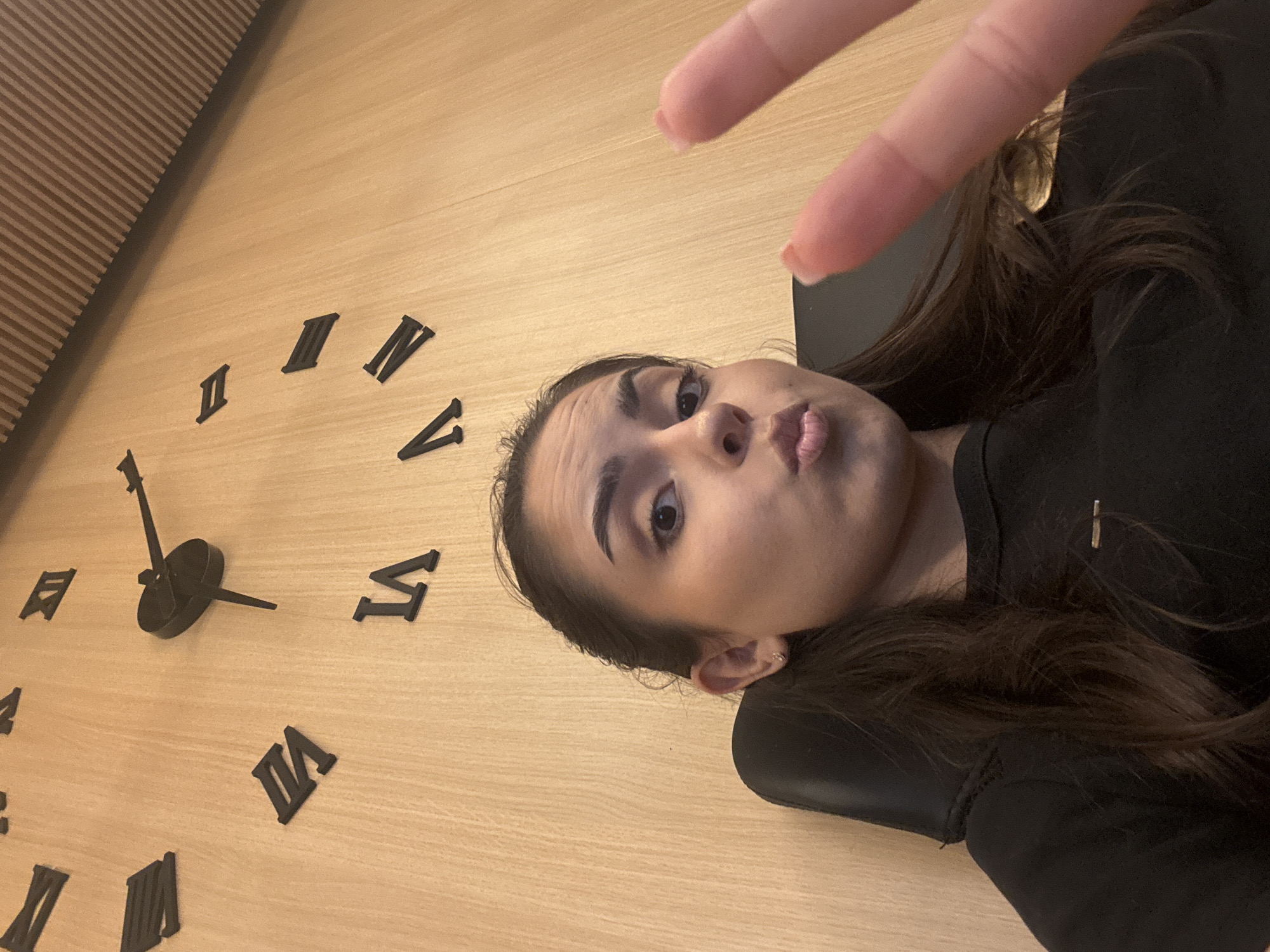 A woman is taking a selfie in front of a large wall clock with Roman numerals. The clock is mounted on a wooden panel wall. The woman is wearing a black t-shirt and has her hair pulled back. She is making a kissing face and holding up two fingers in a peace sign. The image does not contain enough information to determine the location in the city of Sofia, Bulgaria.