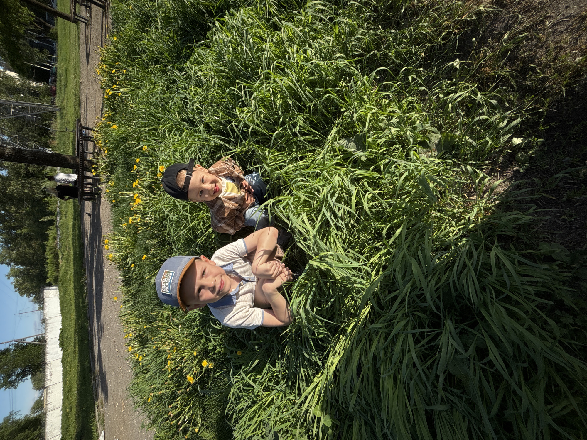 Two young boys are sitting in tall grass with yellow flowers. One boy wears a blue baseball cap and a light-colored short-sleeved shirt, and has his legs crossed and arms around his knees. The other boy wears a black baseball cap, a yellow shirt, and a plaid jacket. They are surrounded by tall green grass and yellow flowers. The background shows a path, benches, and buildings. The location is near the city of Rusanovo, Russia.