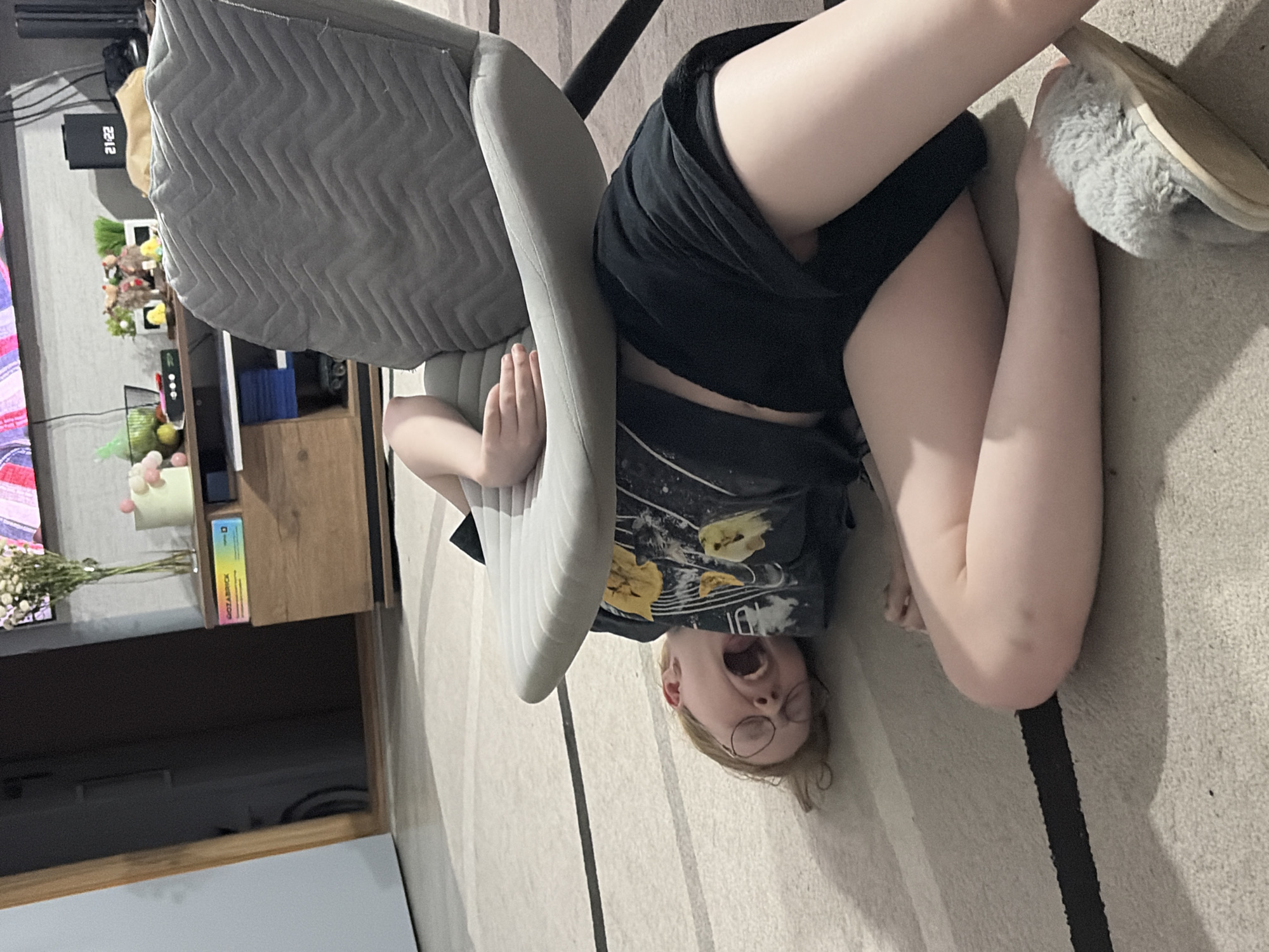 A young person is upside down on a carpeted floor, head near a chair. The person wears glasses, a t-shirt with yellow designs, and black shorts. A fuzzy slipper is visible near their legs. A gray chair is positioned over the person. The setting appears to be a living room or similar interior space, with a television stand and items on it visible in the background. The location is Unknown, Unknown.