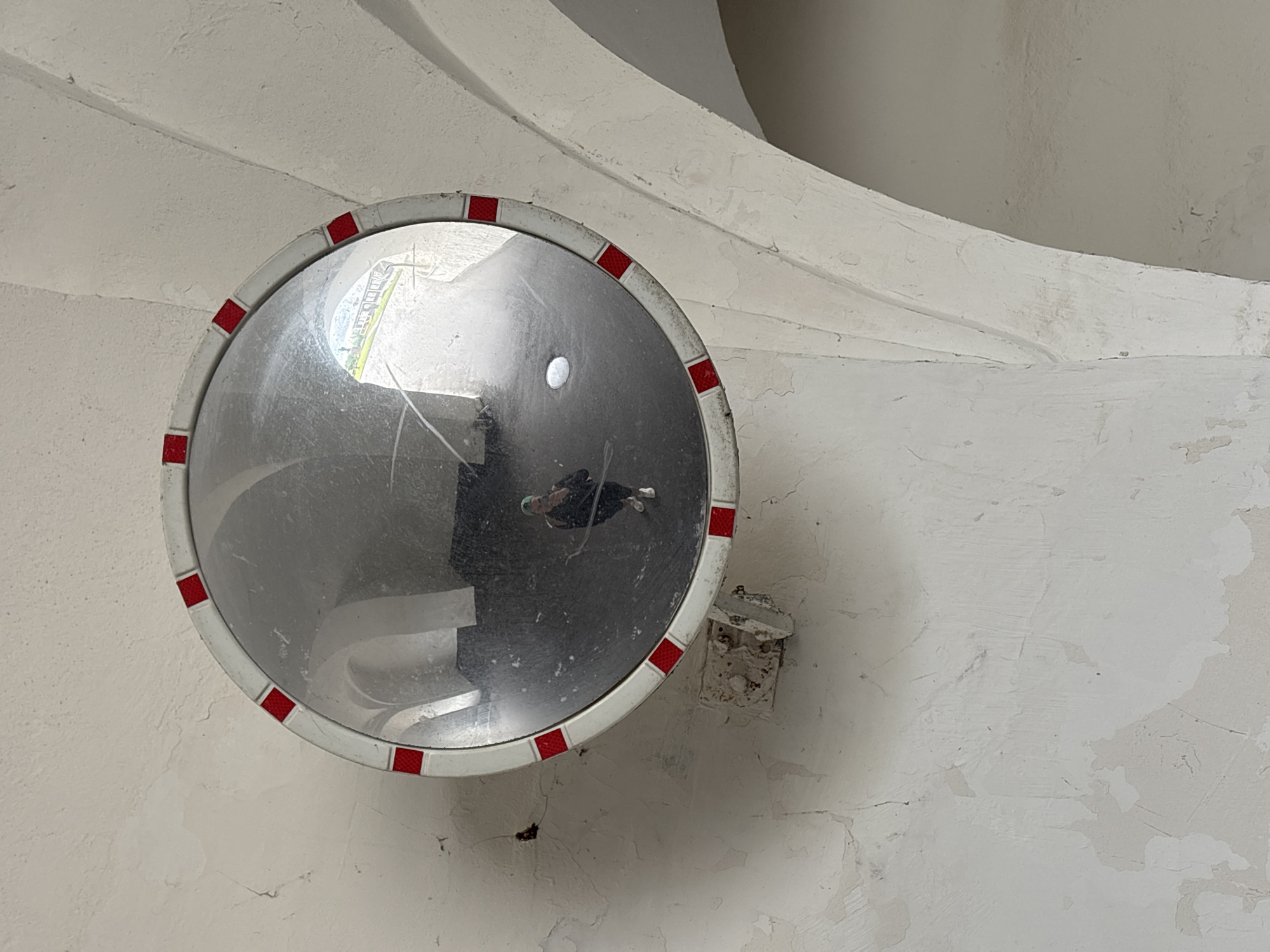A convex security mirror with a white and red-striped frame is mounted on a textured white wall. A person is visible as a reflection in the mirror, holding a phone and taking a picture. The wall appears aged and cracked, with archways visible in the background. The scene is indoors, likely in a covered walkway or building with arched structures. The location is identified as Sof'ino, Russia.