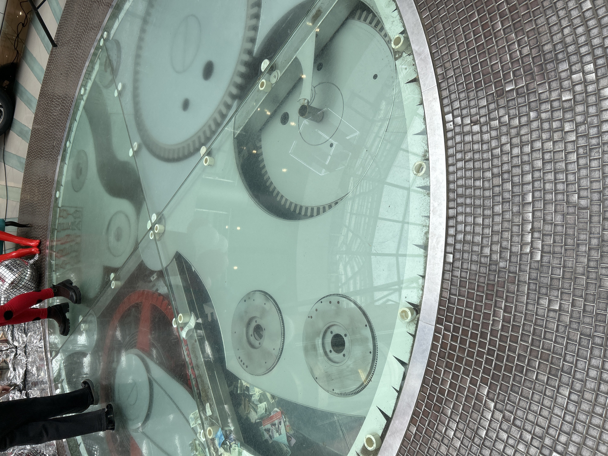 The image shows a circular glass floor revealing a collection of gears, presumably part of a mechanical installation, situated in Izhevsk, Russia. The floor is bordered by a metallic frame. The gears are various sizes and colors, including white, black, and red. Below the glass are items like paper and what appears to be scattered trash. On the glass floor are two people. One is wearing black pants and boots, while the other is wearing red pants and black boots. A metallic, reflective object is next to them. The floor surface adjacent to the glass is tiled with a small square grid pattern.
