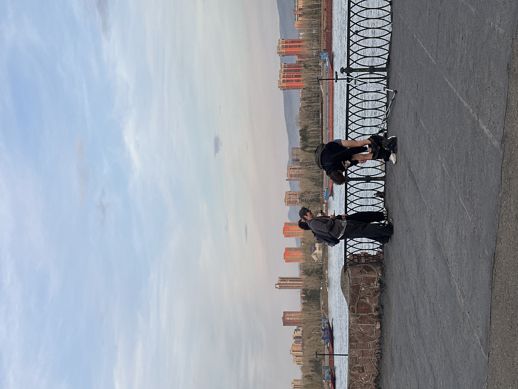 The image shows a waterfront scene in Krasnoyarsk, Russia. In the foreground, two people are standing next to a decorative metal railing. One person is bent over, possibly stretching or tying shoes. The other person is standing and appears to be looking out at the water. Behind them, the Yenisey River flows past a backdrop of buildings and trees. The sky is partly cloudy. A barge is docked on the waterfront.