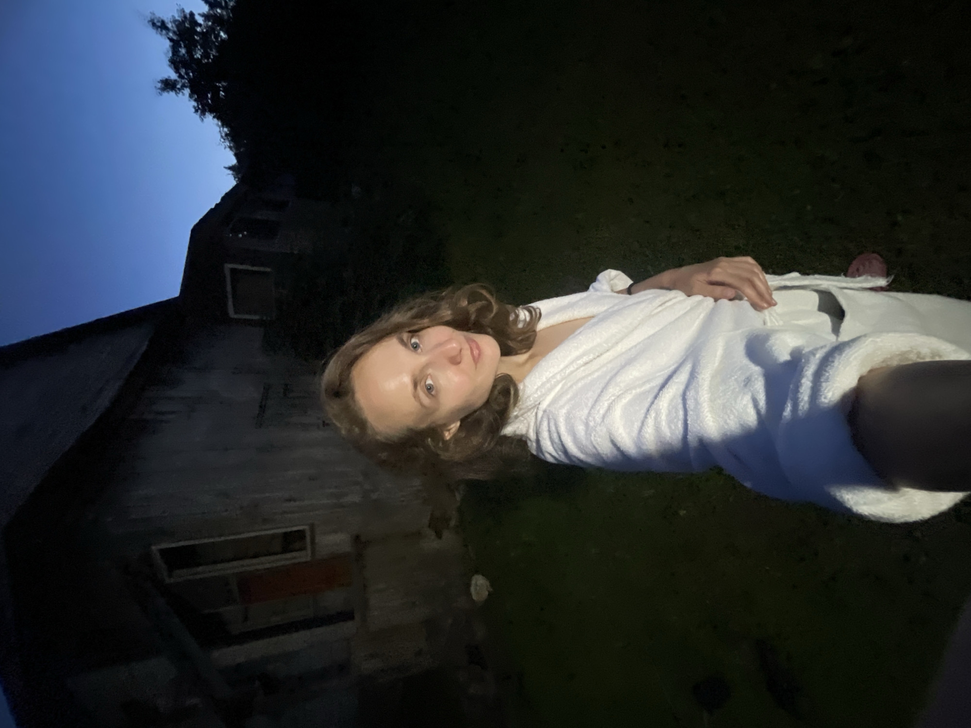 A woman with wavy brown hair, wearing a white bathrobe, stands in front of a dark, weathered building at dusk. The building appears to be abandoned, with boarded-up windows and peeling paint. The sky above is a deep twilight blue, fading to black as it reaches the horizon. Trees and foliage are silhouetted against the darkening sky. The ground is covered in grass, with some patches appearing darker and more shadowed than others. The woman is holding a phone or camera in her right hand, taking a selfie. Her expression is neutral, and her gaze is directed towards the camera. The overall scene suggests a rural or semi-rural setting, possibly in the outskirts of Sharkan, Russia, given the presence of a rustic, older building and natural surroundings.