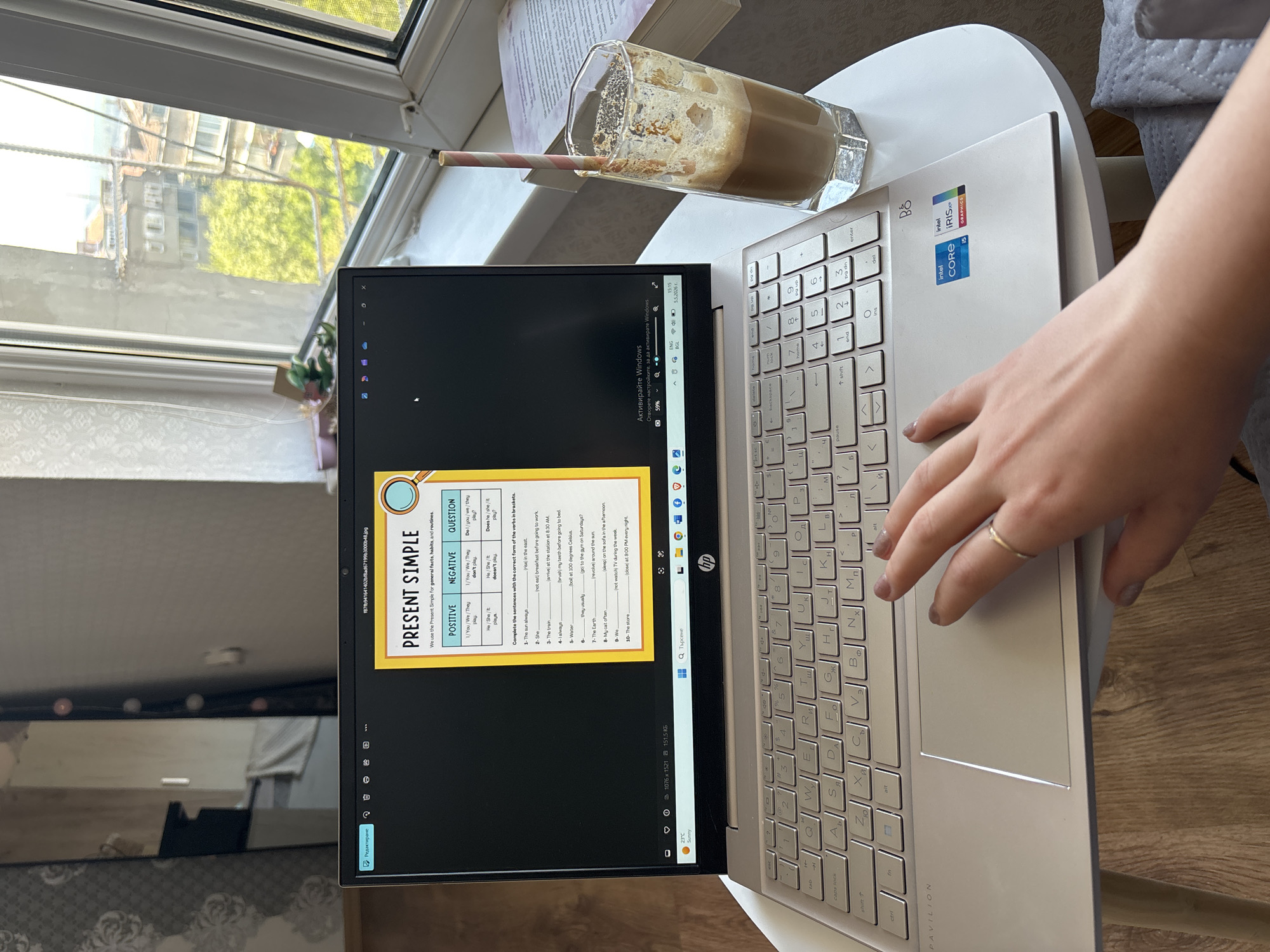 The image shows a laptop displaying an English grammar worksheet on "Present Simple" tense. A right hand with a ring is touching the trackpad of the laptop. Next to the laptop is a partially consumed glass of coffee with a straw. There is a book behind the glass. The laptop rests on a small, round, white table. Through a window, blurred buildings and green trees suggest an urban environment with natural elements. The floor is wooden. The room seems to be an apartment.