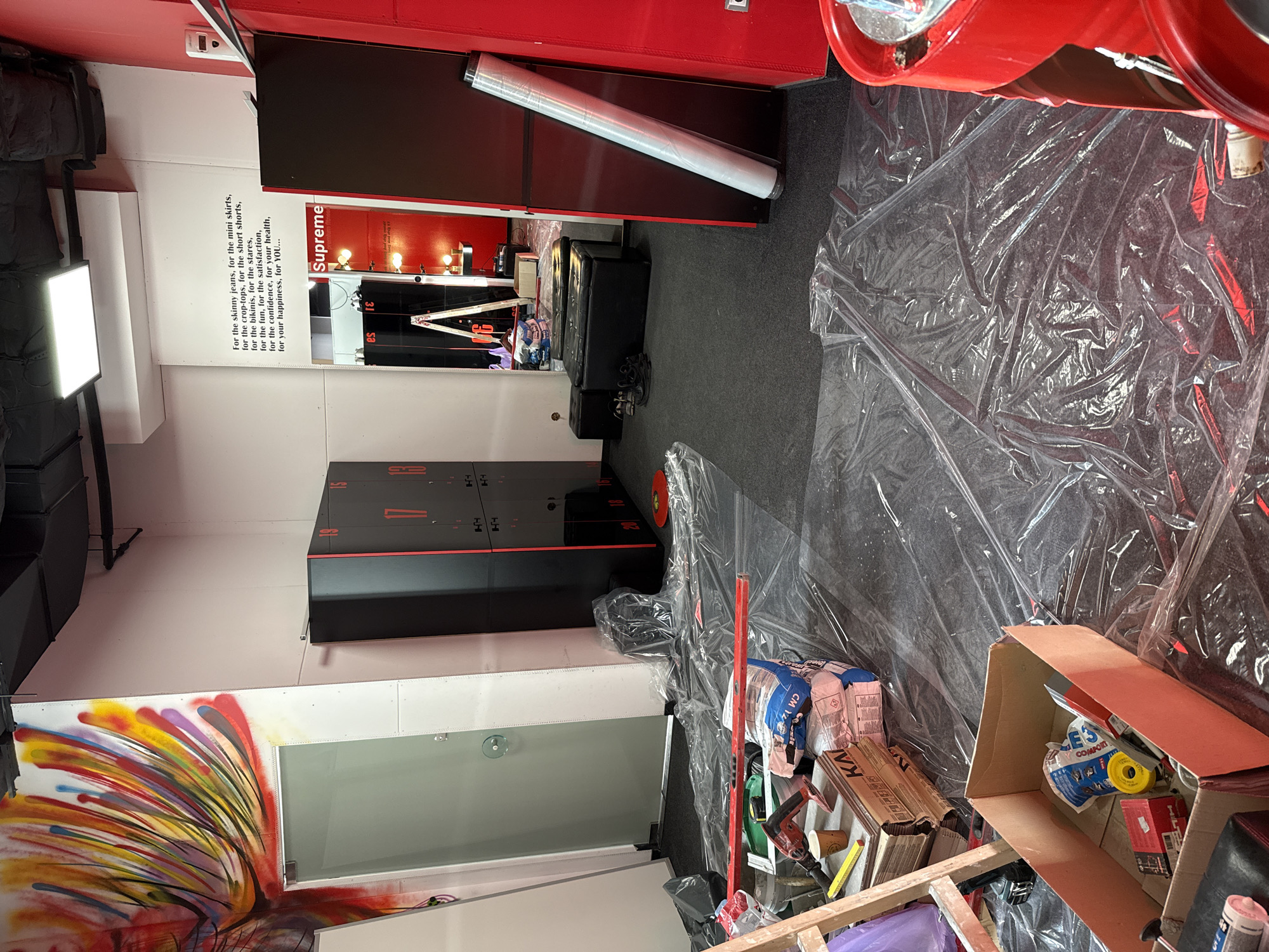 This image captures an interior space under apparent renovation or construction. In the lower left is a partial view of a wall with a colorful, abstract mural resembling an explosion of paint streaks. Above this is a glass partition that appears to lead to another room.
The area is cluttered with materials and objects associated with construction, including opened boxes filled with tools and supplies. Rolls of clear plastic sheeting are strewn across the floor, partially covering objects and the grey carpeting. To the left, a black shelving unit with numbered red labels is visible. Higher up, a horizontal metal bar, possibly a railing or support structure, stretches across.
The background includes text along the wall, which is partially obscured by shadows, but includes the word "Supreme" in a prominent red box. The location is unknown.