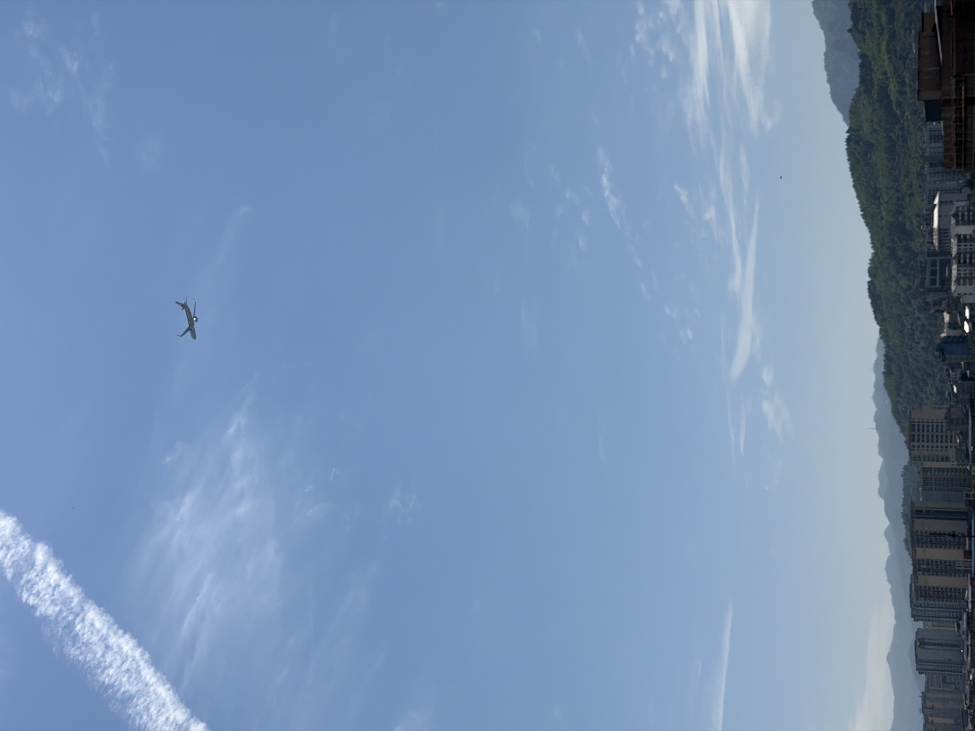 The image shows a plane flying in the sky over Zhangjiajie, China. The plane is in the lower left quadrant of the image and is leaving a contrail. The sky is blue with some wispy clouds. In the right side of the image are buildings and mountains.