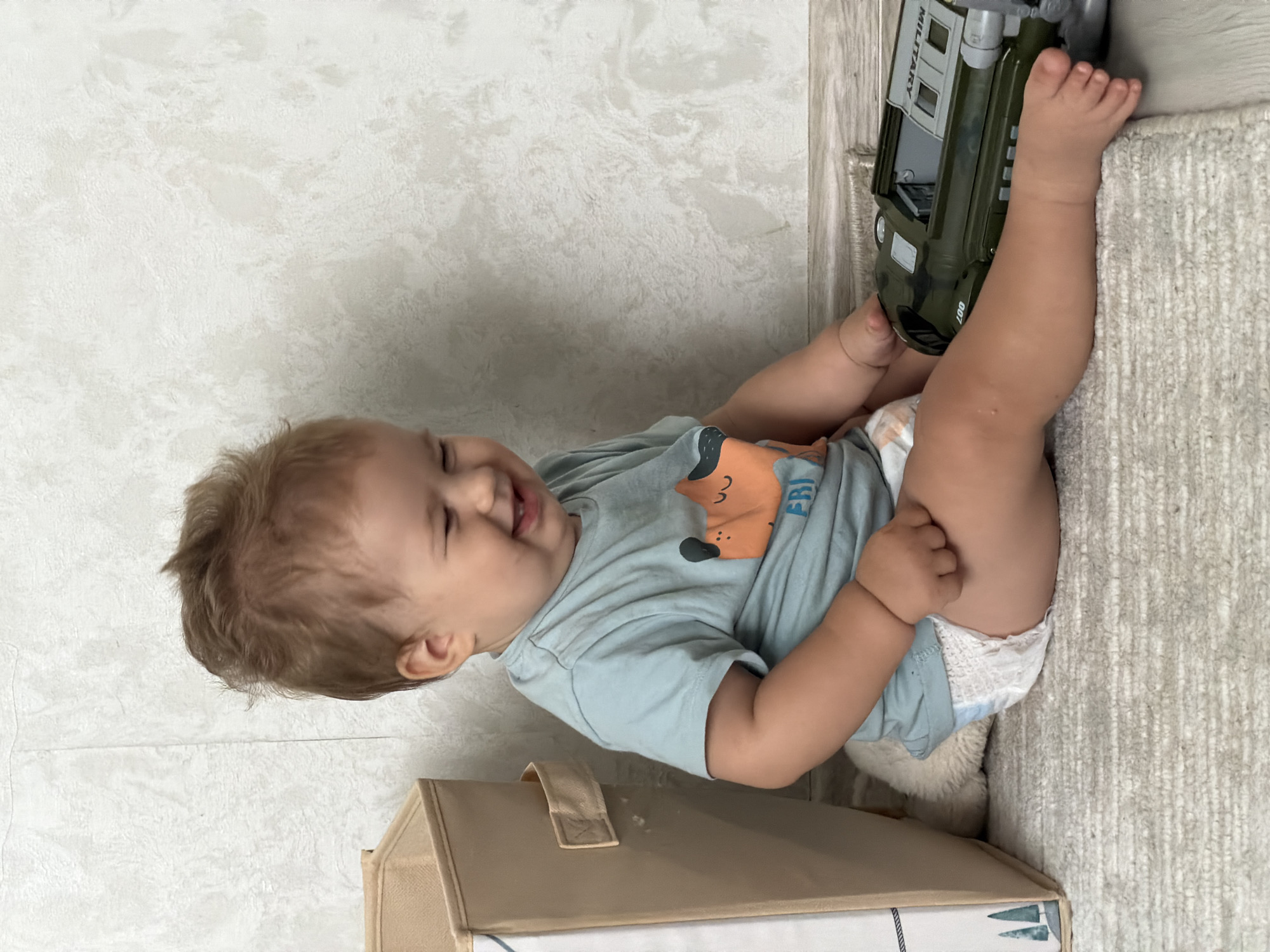 An infant sits on a textured rug next to a fabric storage bin and a green toy vehicle. The infant, with light brown hair, wears a light blue t-shirt with an orange dog graphic and a diaper. The child is laughing and looking to their right. The toy vehicle appears to be a military-style transport with the word "MILITARY" visible. The background consists of a textured, light-colored wall. The scene suggests an indoor domestic setting, likely a child's play area. There are no discernible clues to pinpoint the location to Kazan, Russia.