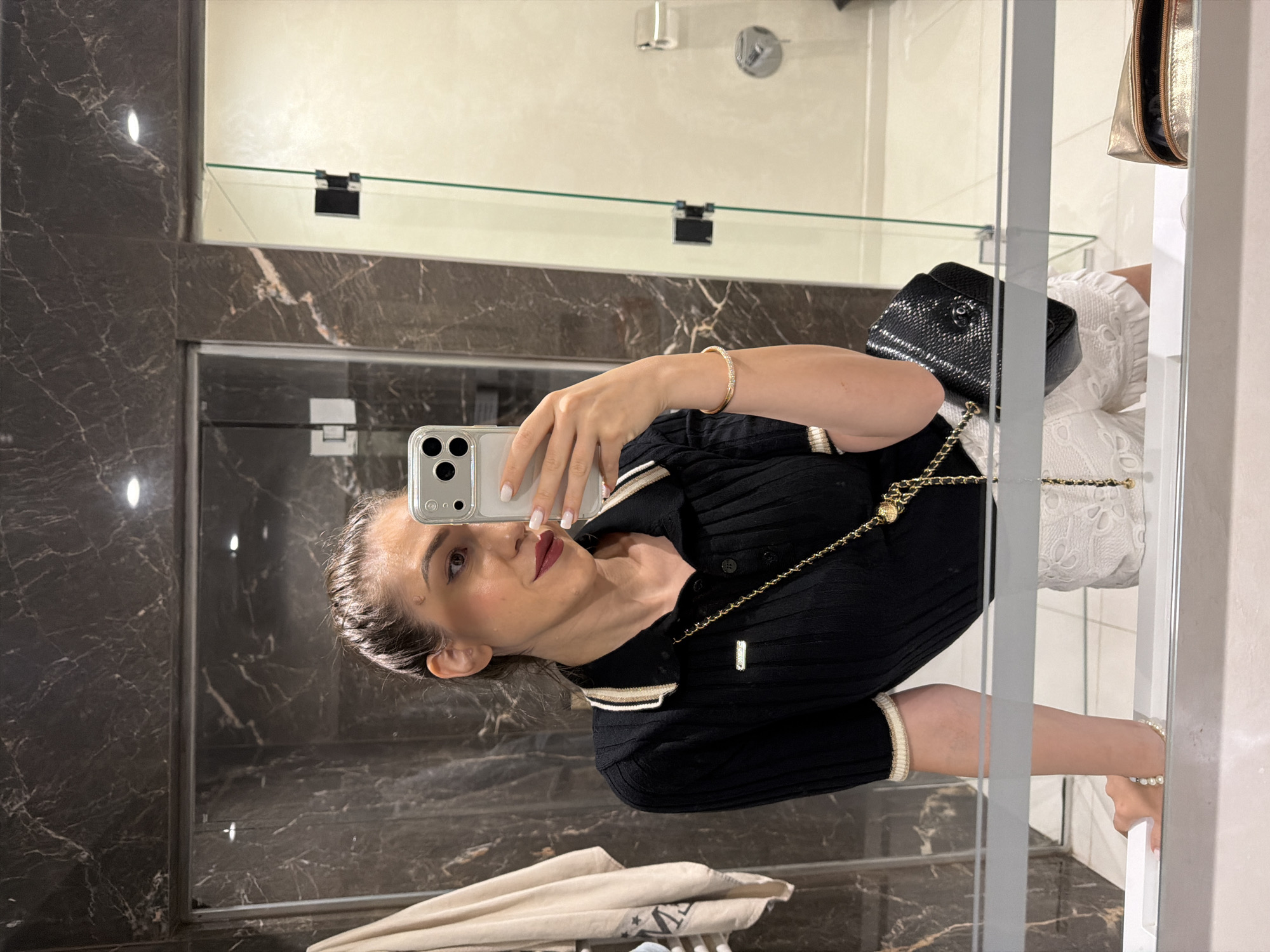 The image is a selfie taken in a bathroom, showing a woman with dark hair pulled back and wearing dark lipstick. She is dressed in a black, short-sleeved, collared top with a ribbed texture and striped trim on the collar and cuffs. A gold chain with a medallion detail is draped across her body, attached to a small black textured handbag. She is also wearing a thin, sparkling bracelet on her wrist.
The background appears to be a modern bathroom with dark marble-patterned walls and a glass shower enclosure. A shelf is visible in the mirror, with a gold cosmetic bag placed on it. The lighting suggests it might be daytime, possibly indoors with artificial lighting. The woman is holding a smartphone to take the photo. There is no visible text in the image.
