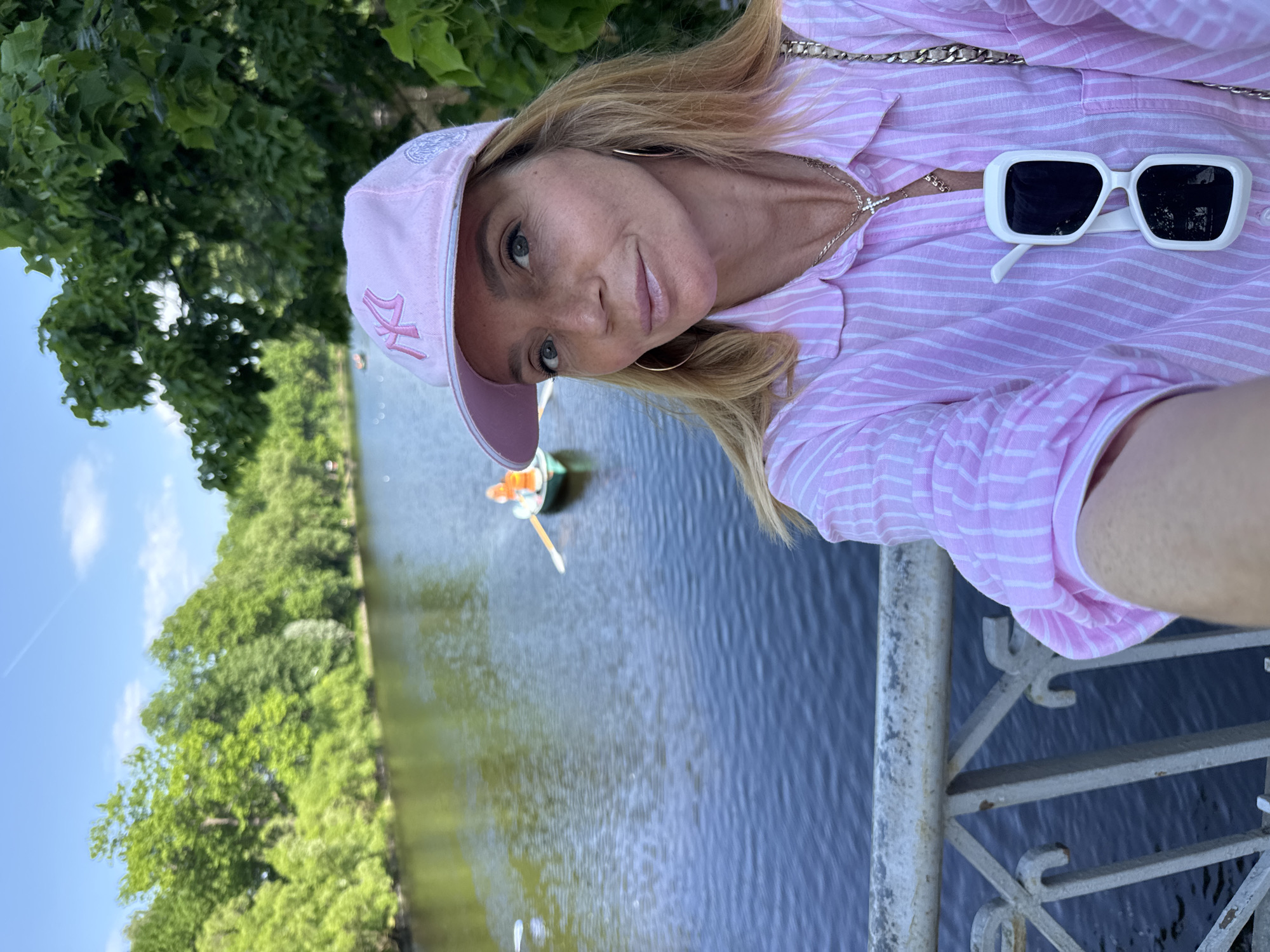 A woman with blonde hair and a pink pinstripe shirt takes a selfie on a bridge overlooking a body of water. She is wearing a pink baseball cap with a New York Yankees logo and gold hoop earrings. A pair of white sunglasses is hanging from her shirt. In the background, a small boat with two people is rowing on the water, surrounded by lush green trees. The sky is blue with a few wispy clouds. The railing of the bridge is visible in the foreground.