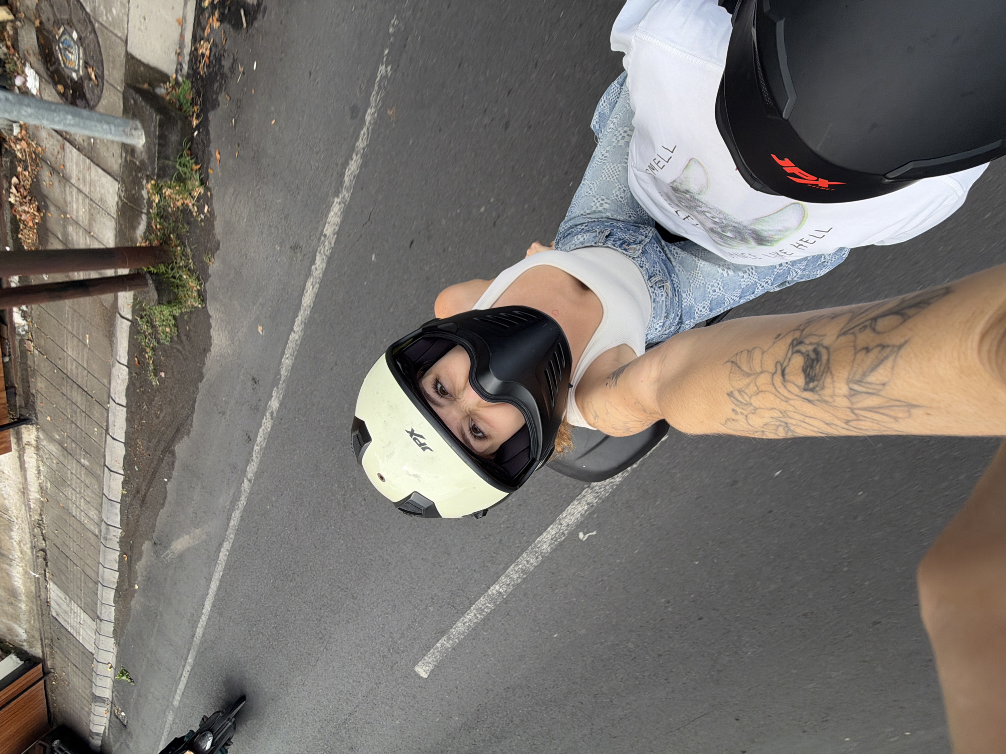 A person wearing a white tank top, denim jeans, a light yellow helmet with black visor, and black knee pads rides on the back of a motorcycle. The person has tattoos on their left arm and a small heart tattoo on their chest. The motorcycle is on a paved road with a white line. A second motorcycle is visible in the background on the left side of the image. The environment appears to be a city street with a sidewalk and some vegetation. The scene is captured from a high-angle perspective, looking down at the rider and the road. The image was taken in North Kuta, Indonesia, as indicated by the presence of a street sign with "Jalan Raya Canggu" and the overall urban environment.