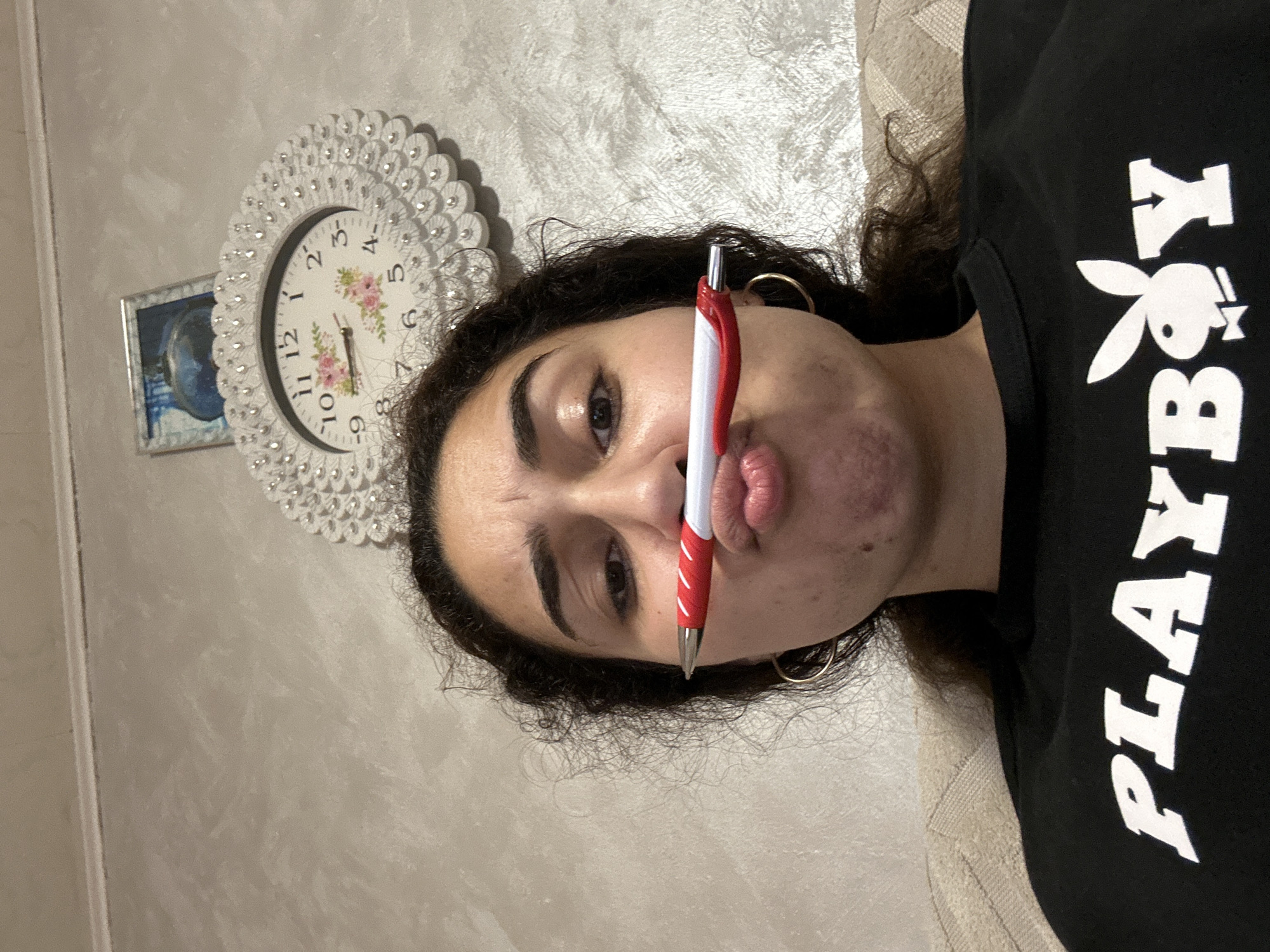 A woman lies on a beige upholstered surface. She is holding a red and white pen in her mouth. She is wearing a black t-shirt with white "PLAYBOY" lettering and the iconic bunny logo. She has dark curly hair, dark eyes, and is wearing gold hoop earrings. Behind her on a beige wall is a decorative white clock with a floral design and a framed image. No information is available to verify whether this image was taken in the city of Burgas, Bulgaria.
