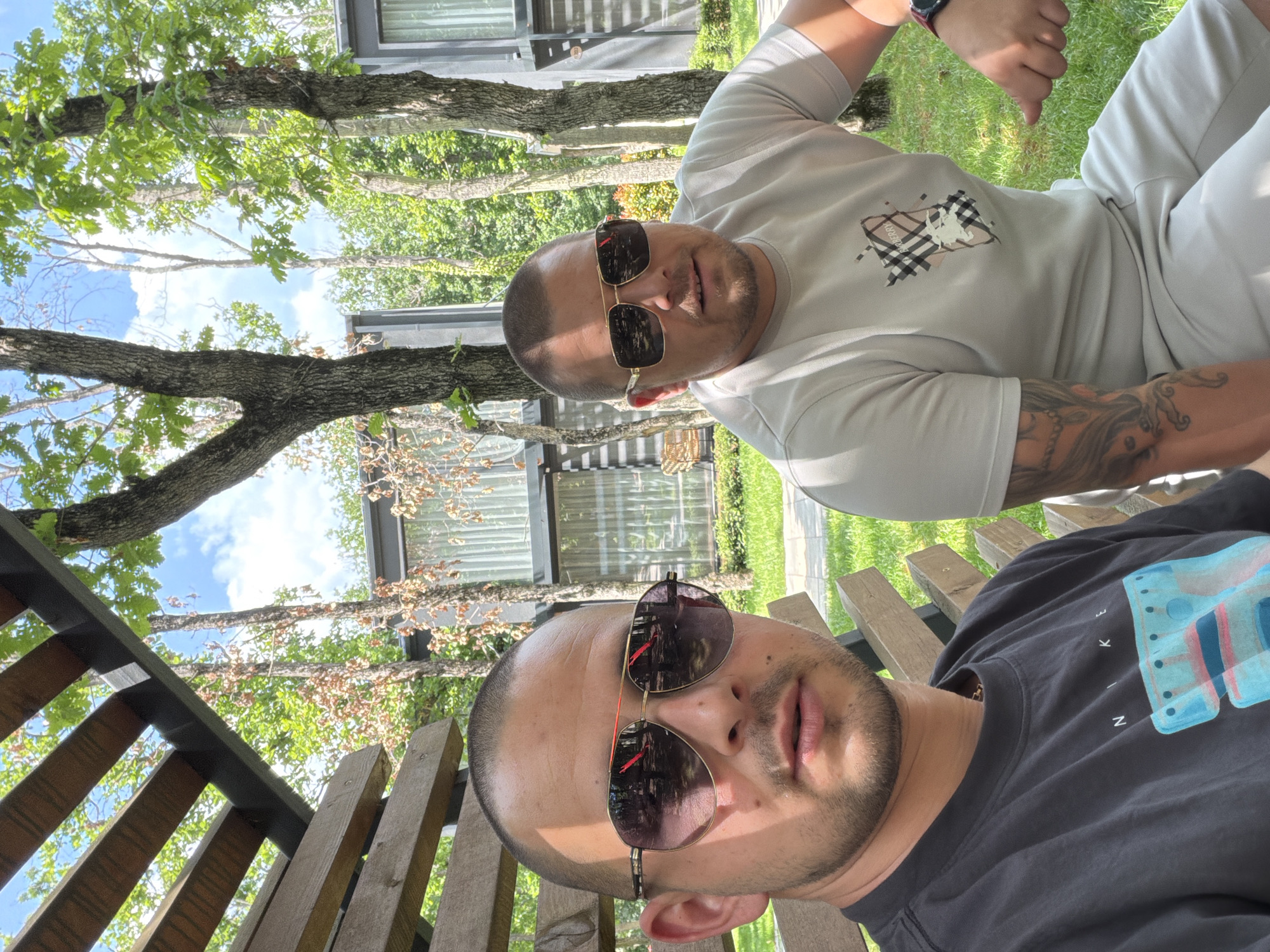 Two men wearing sunglasses sit outside on a wooden bench or deck. One man wears a light colored t-shirt with a Burberry design and light colored shorts, and has a tattoo on his arm. The second man wears a dark t-shirt. Trees and a building are visible in the background. The location is possibly Burgas, Bulgaria.
