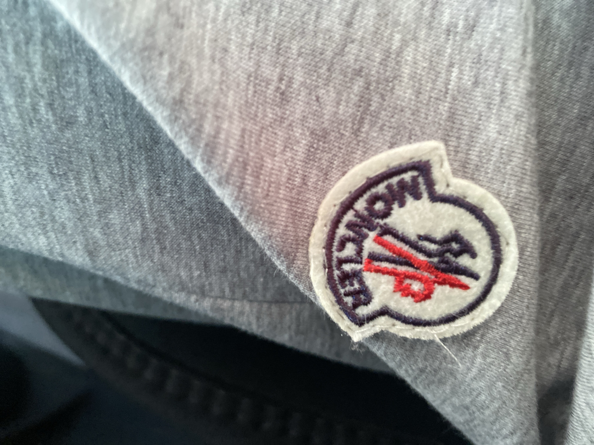 A close-up, overhead shot shows a grey hooded sweatshirt with a Moncler logo patch. The patch is circular with a white background, a dark purple border, and the Moncler logo in red and dark purple in the center. The sweatshirt is laid out, with the hood partially obscuring the background. There are no people or other objects visible in the image. The scene is indoors, with soft lighting.