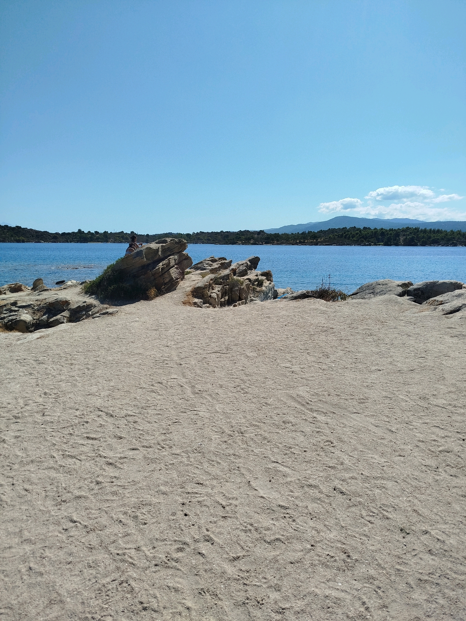 A wide-angle, eye-level shot captures a sandy beach leading to a rocky outcrop, with a calm, blue sea and a treed shoreline in the background under a clear sky. A single person is seated on the rocks on the left side of the frame. The sand is light brown and shows faint footprints. The rocks are beige and irregularly shaped. The water is deep blue and still. The shoreline is lined with dense green trees and vegetation, with faint outlines of hills in the far distance. The sky is a uniform, pale blue. The location is the Sithonia Municipal Unit, Greece.