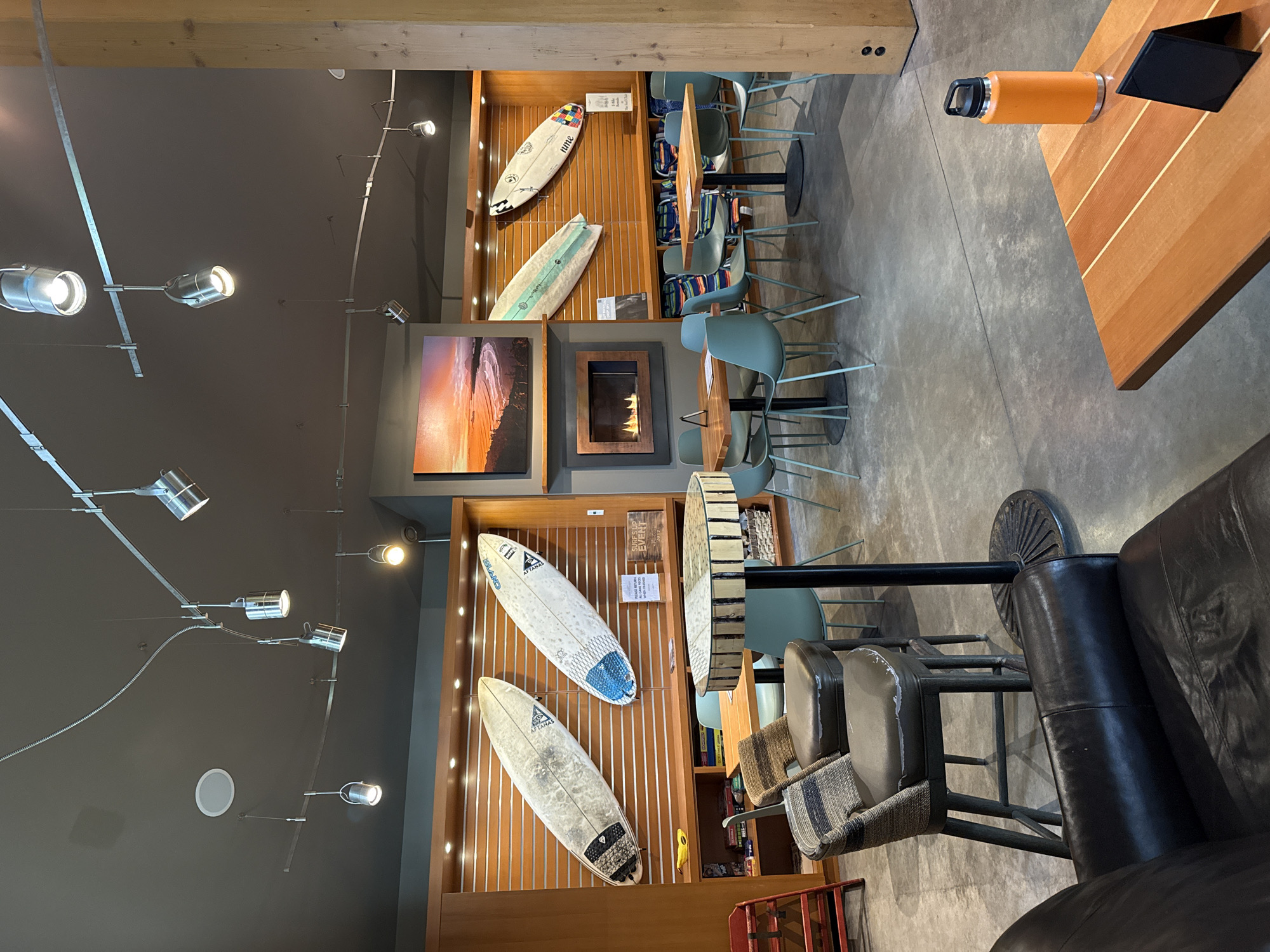 The image depicts an interior space in Tofino, Canada, characterized by a modern, surf-themed aesthetic. A prominent feature is a wall display of surfboards interspersed with decorative wooden slats. Adjacent to the surfboards is a fireplace, above which hangs a landscape photograph. The seating arrangement includes high stools at bar-style tables, and a lounge area with leather seating. Contemporary track lighting illuminates the space. Other objects in the scene include a thermal water bottle, a coaster, and various decorative items on shelves.