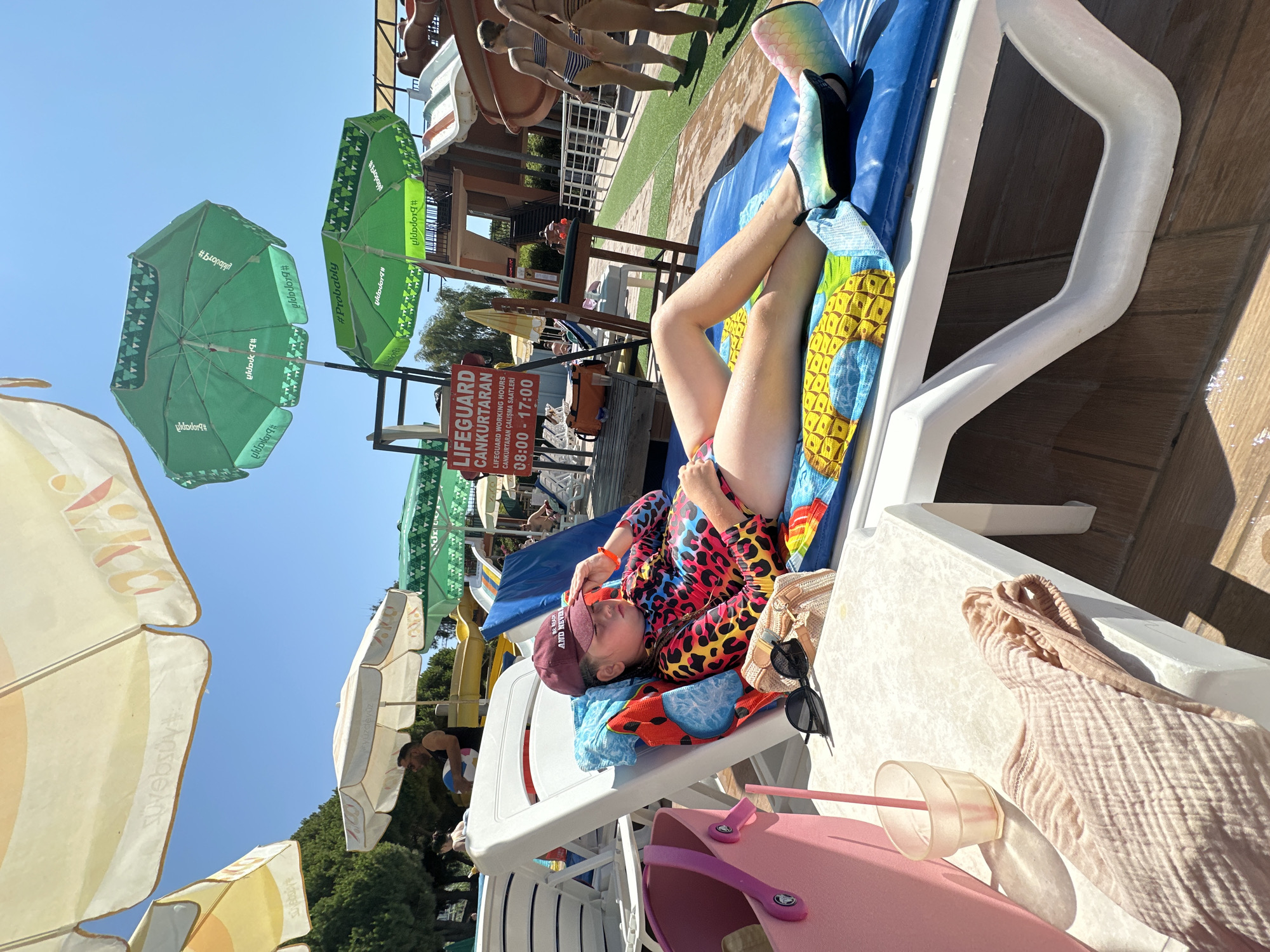 The image captures a sunny day at a water park in Alanya, Turkey. In the foreground, a young girl is lounging on a white plastic sunbed, covered with a blue towel and a colorful towel depicting pineapples. She wears a vibrant, long-sleeved swimsuit with a leopard print pattern and a maroon baseball cap. Her right hand rests on her forehead, and her eyes are closed, suggesting she is shielding herself from the sun. On the sunbed next to her are a pair of black sunglasses and a beige woven bag. To the left of the sunbed is a small white table with a pink cup containing a straw and a folded light-colored towel. In the background, several green and yellow umbrellas provide shade. A red and white sign reads "LIFEGUARD" and lists working hours. Further back, water slides are visible, with a few people walking on the deck. The ground appears to be paved with wooden planks and artificial turf. The sky is clear and blue, indicating a warm, sunny day.