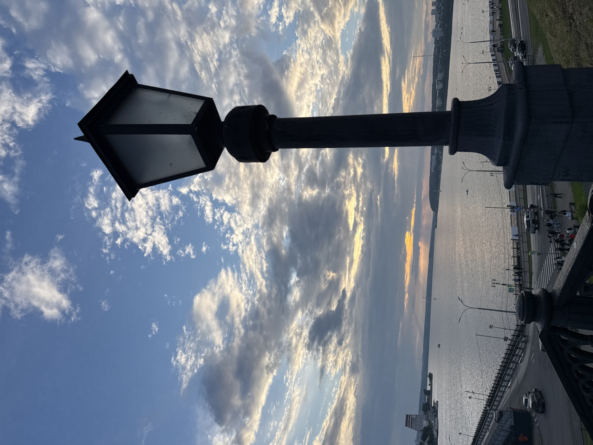 This is a photograph of a river and cityscape, likely taken at dusk. In the foreground, a black lamppost stands prominently, its light diffused by a frosted glass enclosure. Behind it, a wide river occupies the middle ground, with several boats visible on its surface. A bridge crosses the river in the lower left quadrant, with cars and pedestrians present. The far bank of the river features a distant cityscape with buildings and a noticeable industrial structure with red accents. The sky above is filled with dramatic clouds, with rays of sunlight breaking through, suggesting the sun is setting. The scene is located in Izhevsk, Russia, and depicts the Izh River with the Stolyarny Bridge in view.