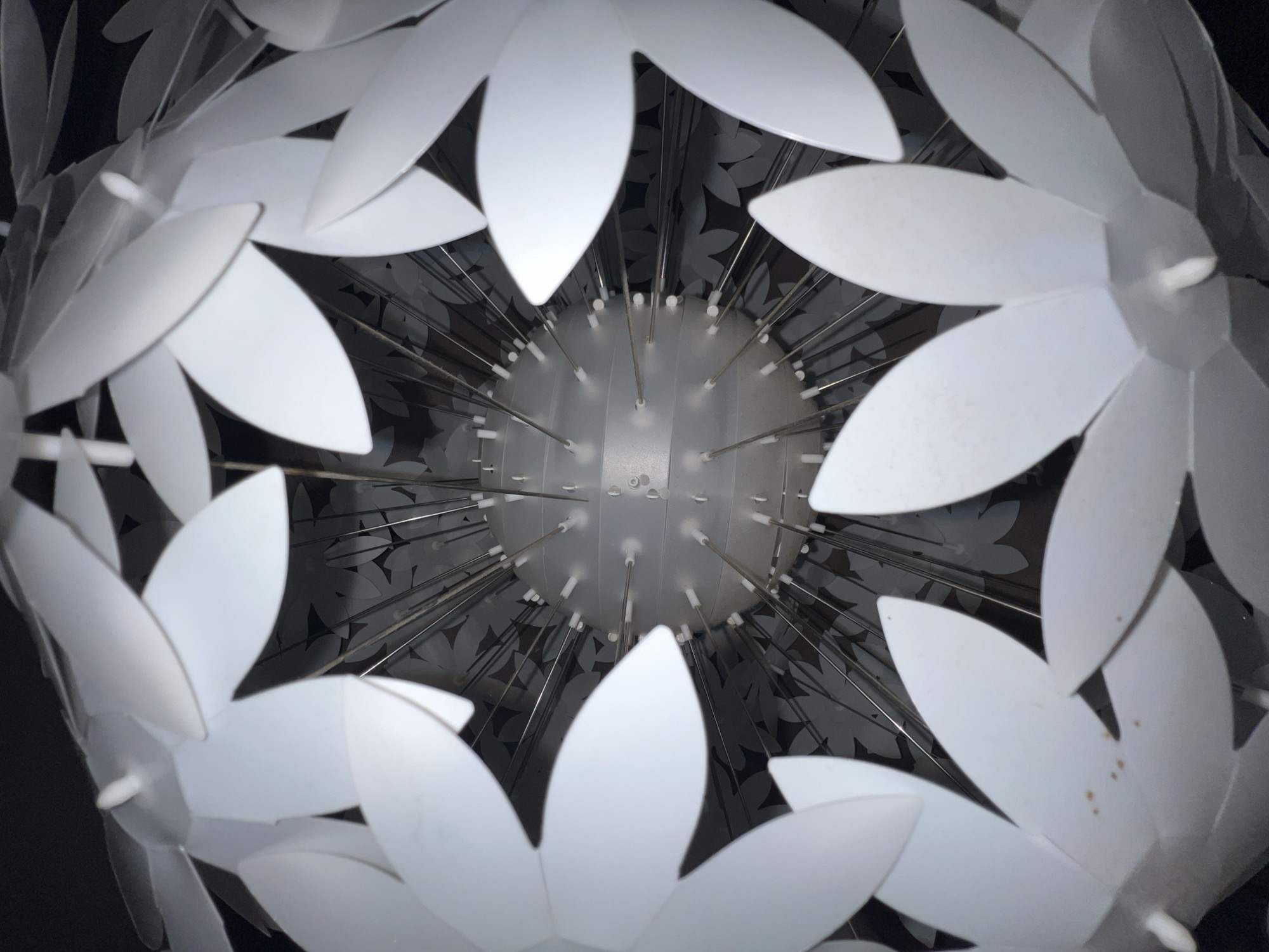 The image is a close-up, downward-facing view of a white, spherical lampshade, possibly made of paper or plastic. The lampshade is composed of numerous petal-like elements arranged in layers. In the center, a translucent spherical core is visible, with thin metal rods extending outwards, supporting the petals. The background is dark and appears to be the interior of the lampshade, showing a repeating pattern of darker flower shapes against a lighter surface. There are no people or actions depicted. The image quality is moderate, with some blurriness and potential artifacts. The location in Madrid, Spain, is not discernible from the visual information provided.