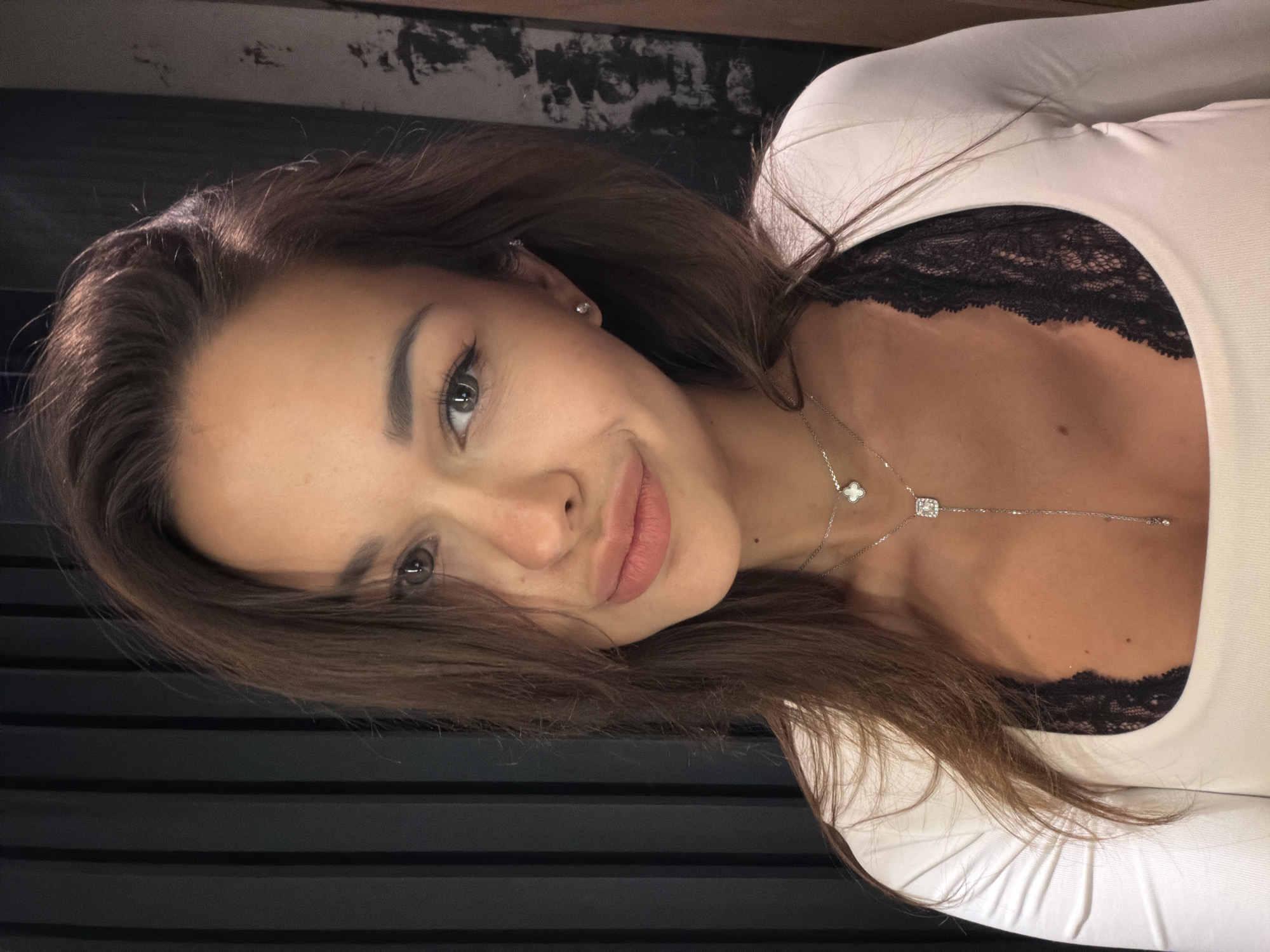 A close-up, waist-up, indoor portrait of a woman with long, brown hair and light skin is captured. She is wearing a white, long-sleeved top over a black lace bra. A delicate silver necklace with a clover charm and a dangling square pendant with a diamond is visible around her neck. The background is dark and textured, with vertical lines on the left and a rough, grimy surface on the right. The lighting is dim, with a harsh shadow falling across the left side of the woman's face. The scene is a dimly lit, interior space, possibly a room in Saint Petersburg, Russia.