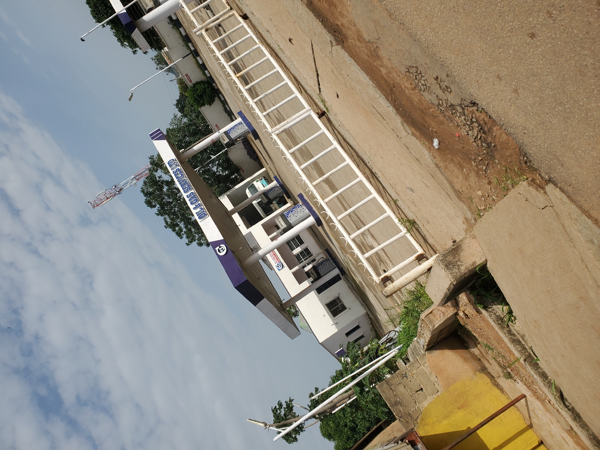 The image depicts an outdoor scene at an oil and gas service station in Bukuru, Nigeria. The primary subject is a white building with a blue and white canopy, characteristic of a gas station. Under the canopy, there are visible gas pumps, one of which has a blue patterned design. A white van is parked near the pumps. The station's name, "OIL & GAS SERVICES LTD," is prominently displayed in blue lettering on the canopy.
To the left of the station, there is a yellow wall with a rough texture, and some green vegetation is visible around the base of the building and the station's infrastructure. A white metal fence runs along the right side of the image, separating the station's forecourt from the viewer's perspective. The ground surface appears to be a mix of concrete and asphalt, with some areas showing wear and tear, including loose gravel and a small puddle of water.
In the background, trees and other vegetation can be seen, along with a tall communication tower with red and white markings. The sky is cloudy, with patches of blue visible. The image is taken from a slightly elevated, angled perspective, with the ground and structures appearing tilted. No people are directly visible in the frame.