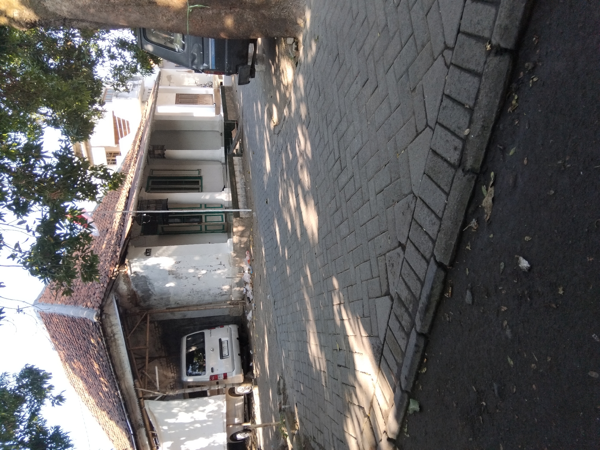 A street scene in Kepuh Kiriman, Indonesia, features a light-colored van parked under a covered carport attached to a weathered building with a tiled roof. The building has a white facade, visible columns, and windows with green trim. Adjacent to the building is a dark-colored vehicle, possibly a pickup truck, partially obscured by a tree trunk. The ground is paved with interlocking gray bricks, and a darker asphalt surface runs along the foreground, littered with some debris. Overhead, tree branches with green leaves cast dappled shadows on the paved area. The sky is visible and appears clear.