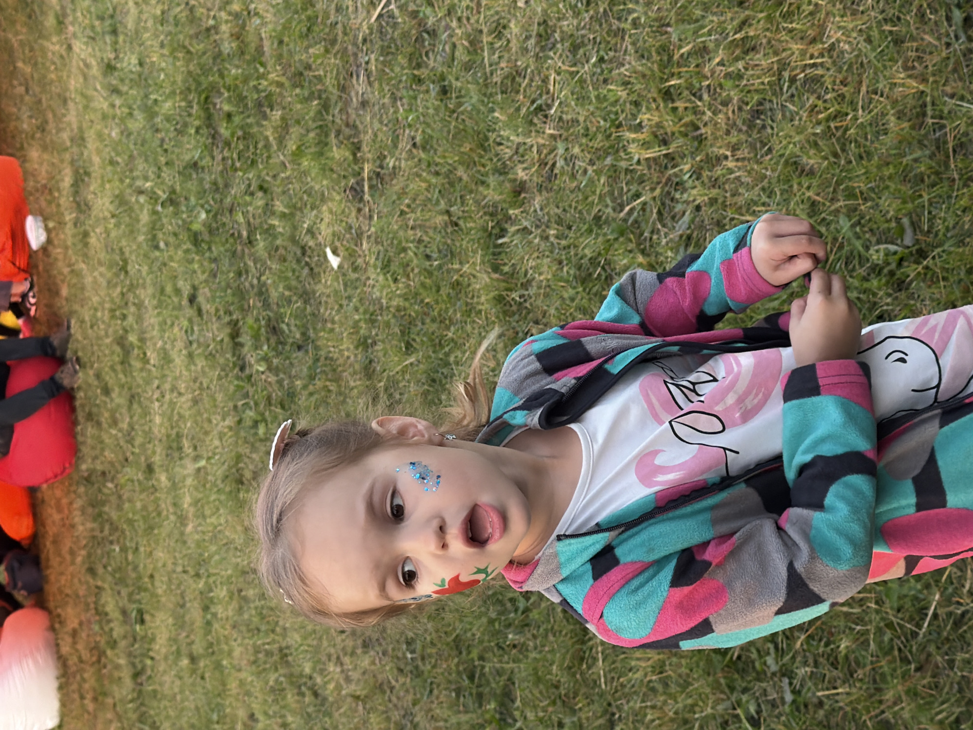 A young girl with face paint stands on a grassy lawn. She is wearing a colorful jacket with a black, pink, turquoise, and grey geometric pattern, over a white t-shirt with a pink flamingo design. The girl has brown hair pulled back in a ponytail and is looking to her left with her mouth open. There is red and green face paint on her left cheek, and blue glitter on her right cheek. In the background, partially visible, are large, colorful beanbag chairs. The scene appears to be outdoors, possibly in a park or backyard.