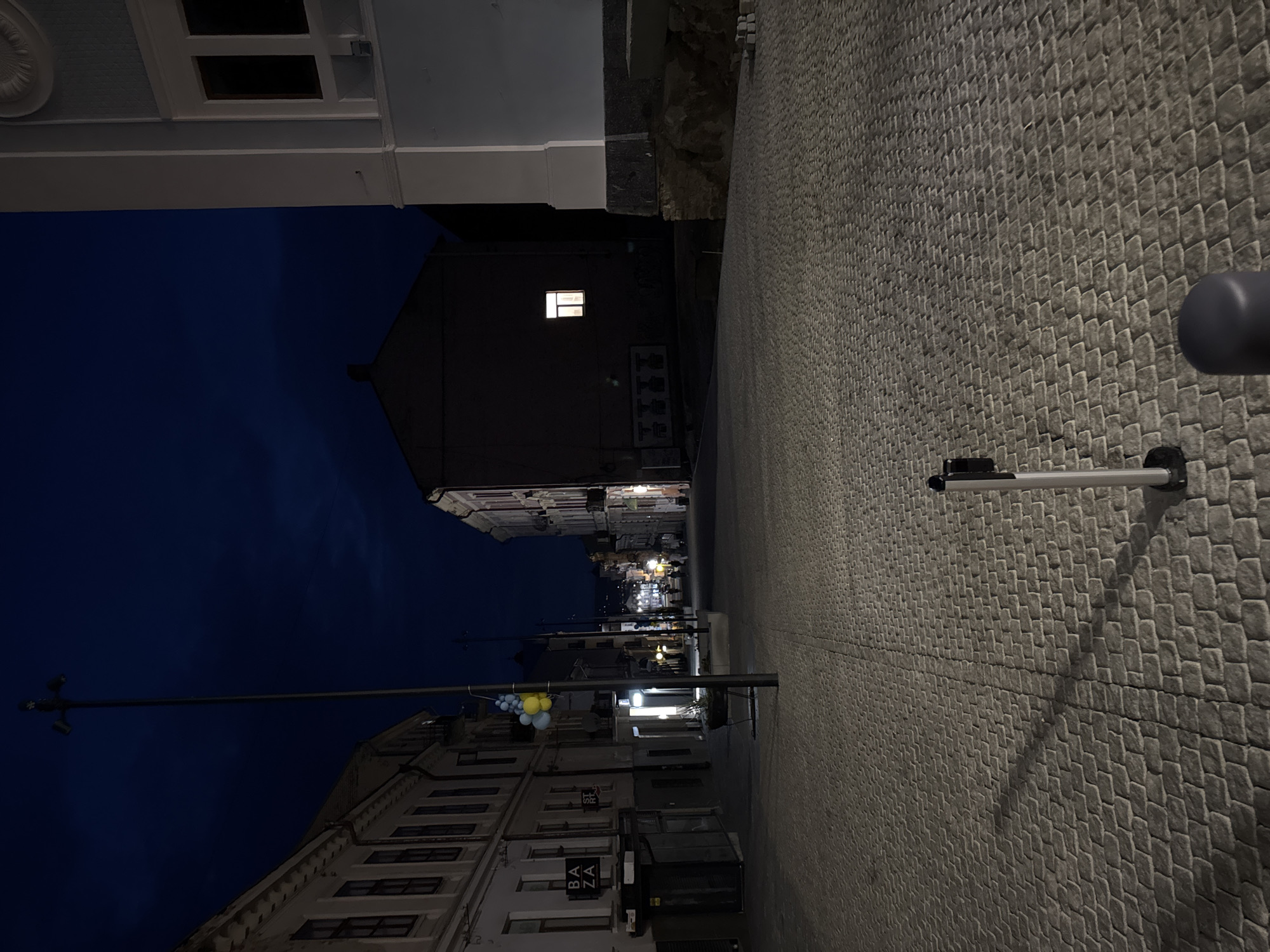 The image depicts an empty cobblestone street at night in Chernivtsi, Ukraine. The street is lined with buildings on both sides, appearing to be a city center or historic district. The sky is dark blue, indicating either late evening or early morning. A tall lamppost with multiple lights stands prominently on the left side of the street, adorned with yellow and blue balloons. In the mid-ground, a building on the right has a single lit window, casting a faint glow. Another building on the left has shop signs that read "BAZA" and "STR T". The cobblestone pavement is well-maintained and reflects the dim ambient light. There are no people visible in the immediate foreground or mid-ground, but a few figures can be seen in the distance, suggesting the street is not entirely deserted. Several bollards are strategically placed along the walkway. The overall atmosphere is quiet and somewhat deserted, typical of a city street after hours.