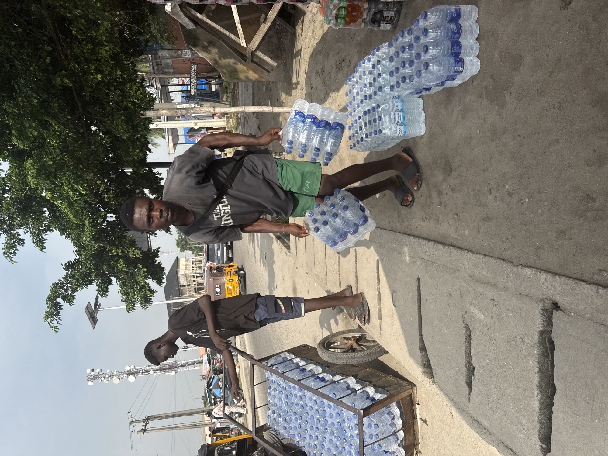 The image captures a bright, sunny outdoor scene depicting two young men engaged in the vending or transportation of bottled water in Ado, Nigeria.
In the foreground, a young man, wearing a dark grey t-shirt, green shorts, and dark sandals, stands on a paved concrete surface. He holds several packs of bottled water, with more packs accumulated at his feet. The water bottles appear to be a brand like "CIDIANIA" or "CIDIANA," visible on the labels. His expression is neutral as he looks towards the camera.
Behind him and to the left, another young man, dressed in a black t-shirt, dark shorts, and sandals, is pushing a metal pushcart. This cart is heavily laden with numerous packs of bottled water, indicating a substantial quantity for sale or transport. An old tire is also visible attached to the cart.
The setting appears to be a street or roadside. The background features lush green trees, various buildings (some appearing unfinished or weathered), and wooden structures that might be market stalls. Utility poles and lines crisscross the scene, and a large communication tower stands tall against the bright, clear sky. A yellow auto-rickshaw (Keke Napep) is partially visible on the street. A sign in the distance partially reads "AFRIATIF" and "FEMME COULEUR." The time of day is clearly daylight, evidenced by the strong shadows and bright illumination. The overall scene conveys a busy, industrious atmosphere.