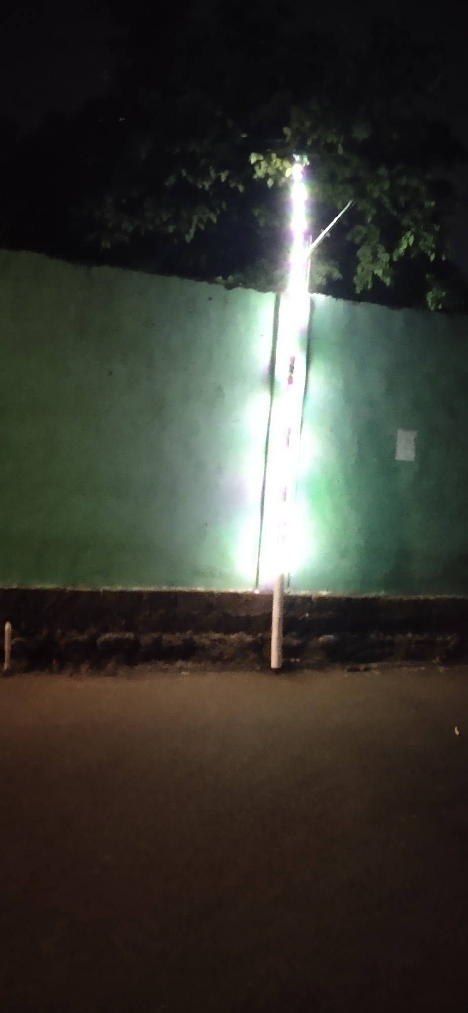This is a night-time, full-shot, eye-level, medium-distance, outdoor photograph taken in Sumengko, Indonesia. The image depicts a tall, slender, illuminated pole with multiple lights running vertically up its length, positioned in front of a solid, green wall. The pole is slightly off-center to the right of the frame. The lights on the pole are a bright white, with a noticeable green glow emanating from them and reflecting onto the green wall. Above the wall, dark tree branches with some illuminated leaves are visible against a dark sky. At the base of the wall, a dark, granular texture is present, likely soil or a similar ground cover, with two small, white vertical posts protruding from it, one on the left side of the frame and another near the illuminated pole on the right. In the foreground, a dark, asphalt-like surface, possibly a road or pathway, fills the bottom portion of the image. The overall scene is dimly lit, with the illuminated pole being the primary light source.
