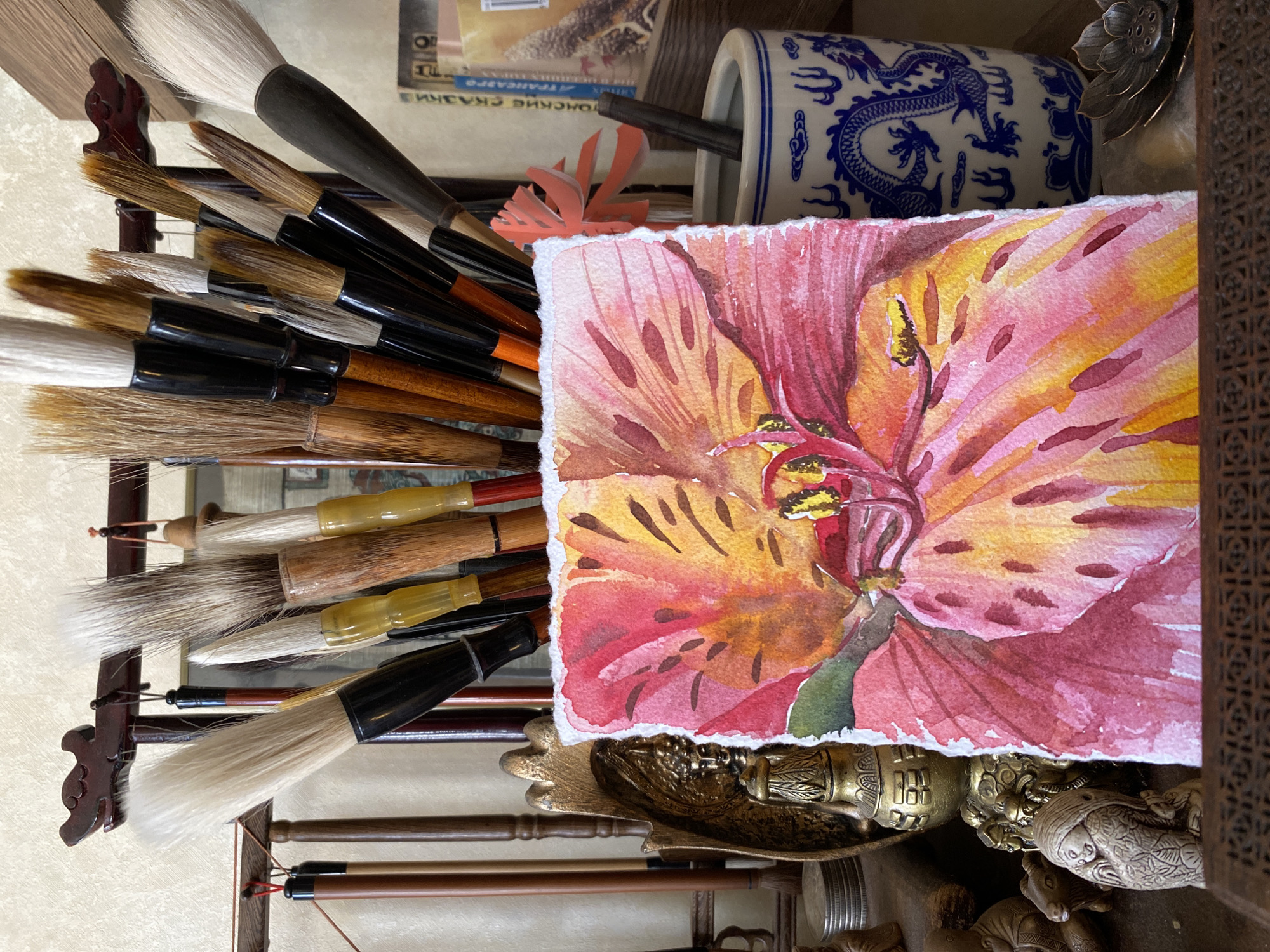 The image depicts a collection of objects arranged in what appears to be an artist's workspace. A variety of calligraphy brushes, with handles in different colors and styles, are bundled together on the left side. A watercolor painting of a flower, primarily in shades of red and yellow, is positioned prominently in the center. Behind the painting, a blue and white ceramic container with a dragon motif is visible. The painting rests on a collection of brass or golden colored objects which appear to be part of the artist's tools. The background is somewhat cluttered. The scene is inside, near a window with natural light. There is no information in the image to suggest the location is Izhevsk, Russia.