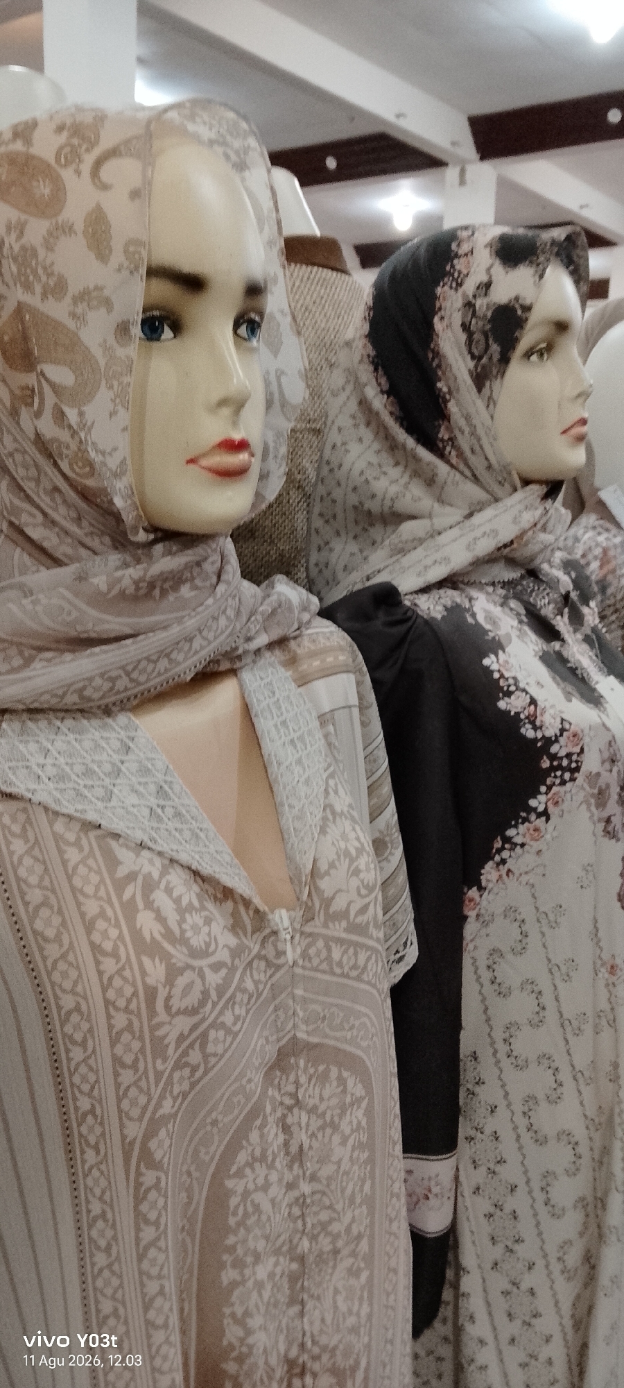 The image displays two mannequins wearing hijabs and dresses, positioned side-by-side. The mannequin on the left is wearing a beige hijab with a paisley print and a beige dress with a floral and paisley pattern. The mannequin on the right is wearing a black hijab with a floral print and a black dress with a white floral pattern. Both mannequins have visible facial features, including blue eyes and red lips on the left mannequin. The background appears to be an indoor retail environment, possibly a clothing store, with soft lighting. A date and time stamp in the lower left corner reads "vivo YO3t 11 Agu 2026, 12.03". The location mentioned is Atula, Indonesia.