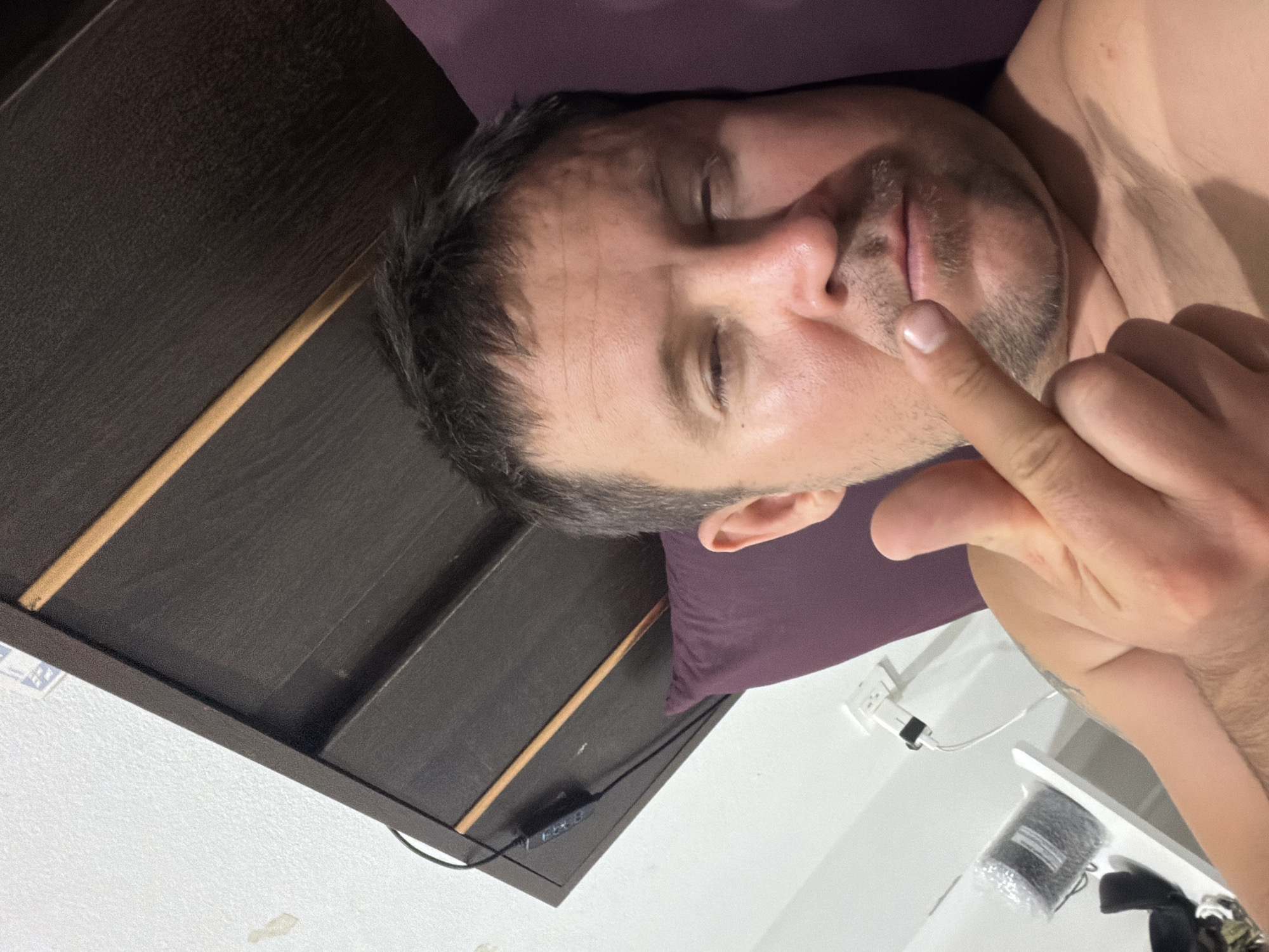 A man is lying in bed, taking a selfie. He is looking at the camera with his eyes partially closed. His right hand is raised, with his middle finger extended towards the camera. He is wearing a purple pillow behind his head and the headboard of the bed is visible in the background. The wall behind him is white. There is an electrical outlet and a phone charger plugged into the wall. The man's arm is hairy. The image appears to be taken in a bedroom in Baan Mae Nam, Thailand.