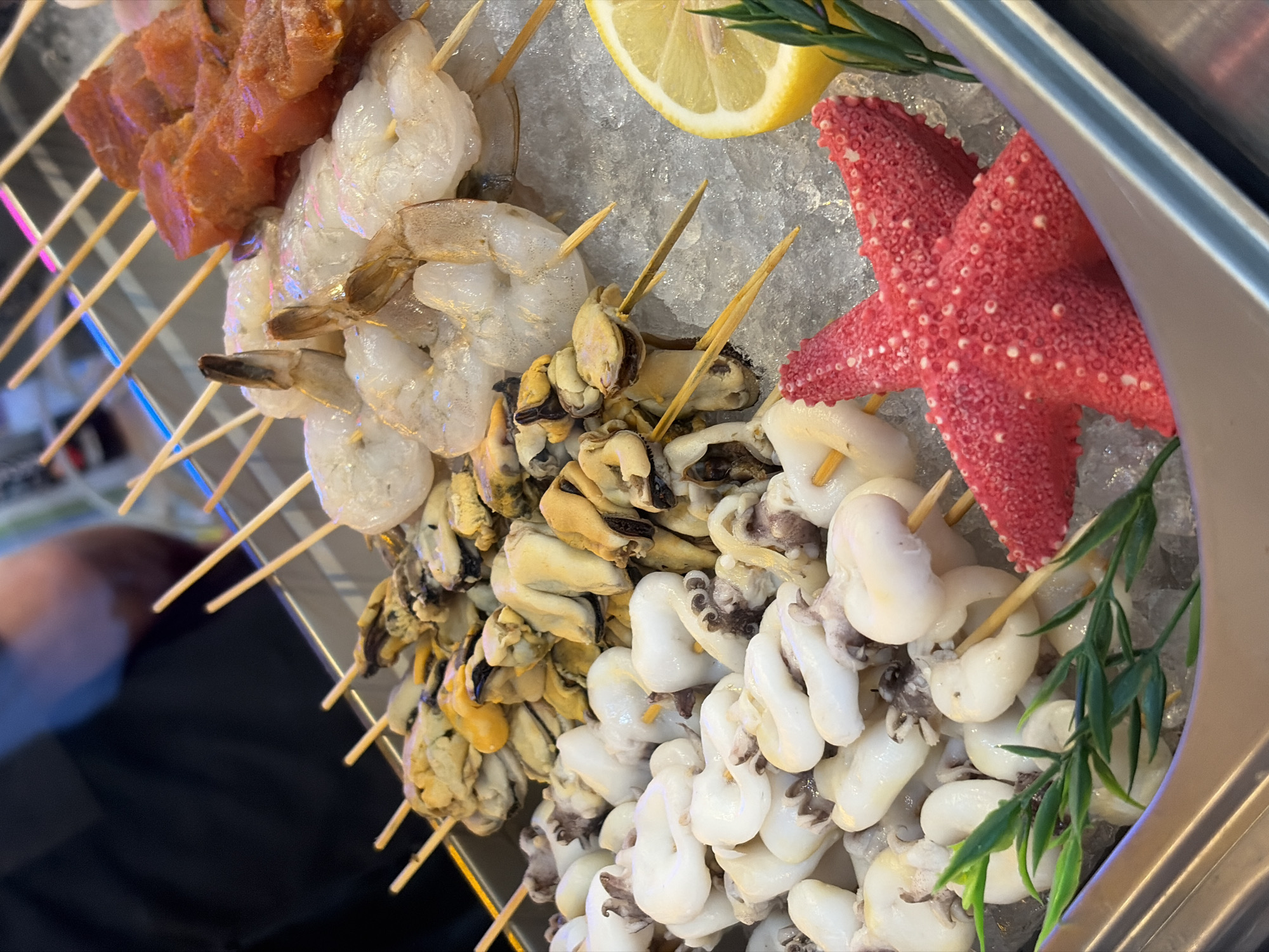 The image captures a close-up, high-angle view of a food display in Saint Petersburg, Russia, showcasing various raw and prepared skewered items arranged on a bed of crushed ice within a silver-toned metal container. In the foreground, a decorative red starfish with a textured surface and a sprig of green artificial foliage are positioned on the ice. To the left, multiple skewers hold small, white, opaque squid bodies, some exhibiting short tentacles. Adjacent to them, skewers display peeled mussels, identifiable by their yellowish-orange flesh. Further right, raw, peeled shrimp with tails intact are skewered, appearing translucent and pale. In the upper right, skewers feature diced, raw red meat, likely pork or beef. A half-slice of yellow lemon is visible on the ice near the shrimp and meat. All food items are presented on thin wooden skewers. The background is out of focus, revealing a person wearing a dark short-sleeved shirt in the upper-left quadrant and indistinct bright lights and reflective surfaces, indicating an indoor commercial food environment.