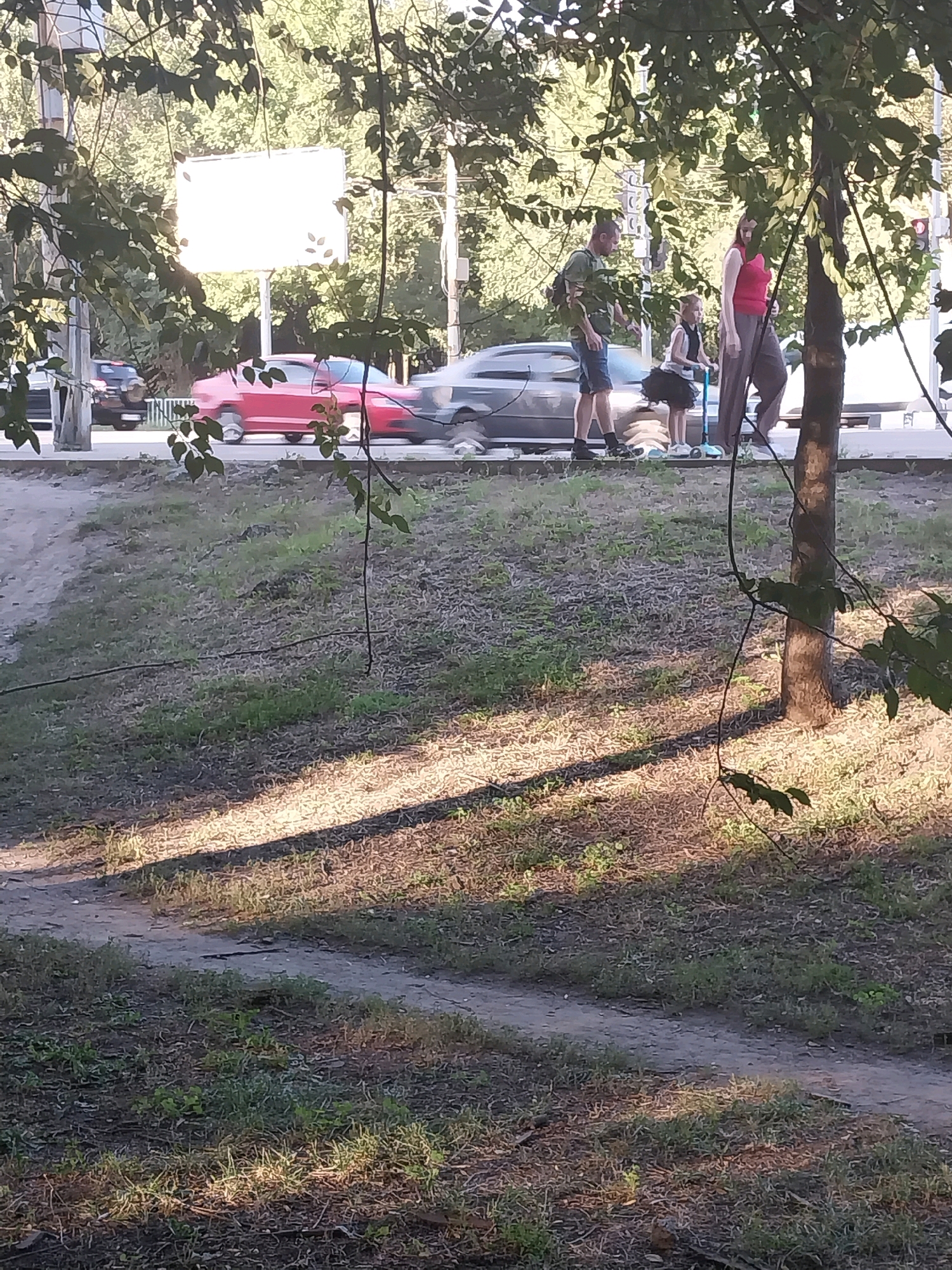 The image shows a street scene in Dnipro, Ukraine, with a man, a woman, and a child near a sidewalk. The man is wearing a green t-shirt and blue shorts, and the woman is wearing a pink top and grey pants. The child is on a blue scooter. In the background, there are cars on the road, including a red car. A large billboard is visible behind the people and cars. The foreground shows a grassy area with shadows cast by trees.