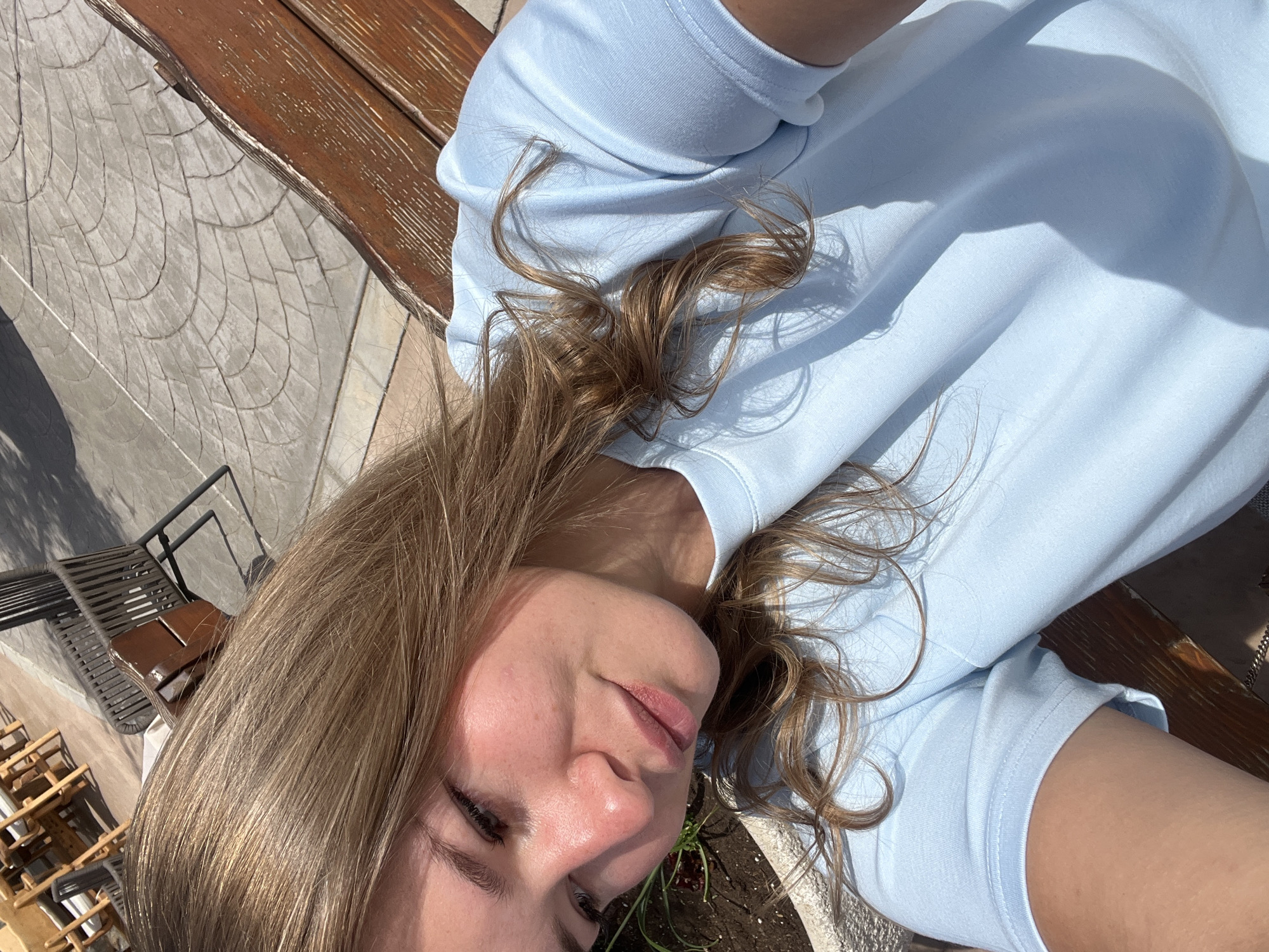 A young woman is captured in a bright, sunlit outdoor selfie, likely taken from an overhead perspective while she is lying down or reclining. Her face, seen from the nose upward, is positioned in the lower right corner, framed by her light brown to blonde hair which spreads across a light blue long-sleeved shirt. She displays a relaxed expression, with a subtle, serene smile.
Strong, distinct shadows fall across her shirt, hair, and the surrounding surfaces, indicating clear skies and direct sunlight, likely during midday or early afternoon. The background elements suggest an outdoor recreational area, possibly a patio or garden in Kovačko Polje, Montenegro. Parts of a dark brown wooden bench or table are visible in the upper left and center right. To the mid-left, a textured grey paved surface with an intricate, scale-like or tiled pattern covers the ground. Further details include sections of dark, woven outdoor furniture, possibly chairs, towards the left background, and a small green plant growing in soil near the woman's face. No visible text is present in the image.