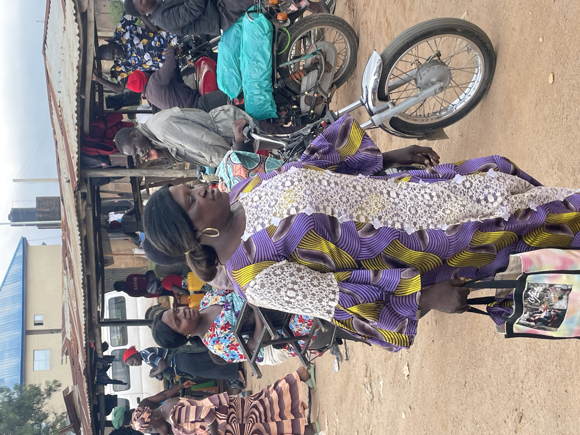 The image depicts a bustling outdoor market scene in Wang, Nigeria, likely during daytime. Several people are present, with a focus on a woman in the foreground wearing a vibrantly patterned purple and yellow dress with white lace detailing. She is looking downwards and holding a tote bag with a colorful tie-dye pattern featuring images. Behind her, another woman in a floral print dress is partially visible, also looking towards the right.
In the background, a motorcycle is prominent, with a turquoise tarp draped over its handlebars. Several other individuals are seen around the motorcycle and a rustic, open-air structure with a corrugated metal roof. One person in a blue and black striped shirt and a red cap is leaning towards the motorcycle. Further back, a white van is parked, and more people are visible, suggesting a busy commercial area. The ground is sandy and dusty, characteristic of an outdoor market. The overall atmosphere appears lively and active, with people engaged in daily activities within the market. No specific time of day or weather conditions are discernible beyond the general brightness of daylight.