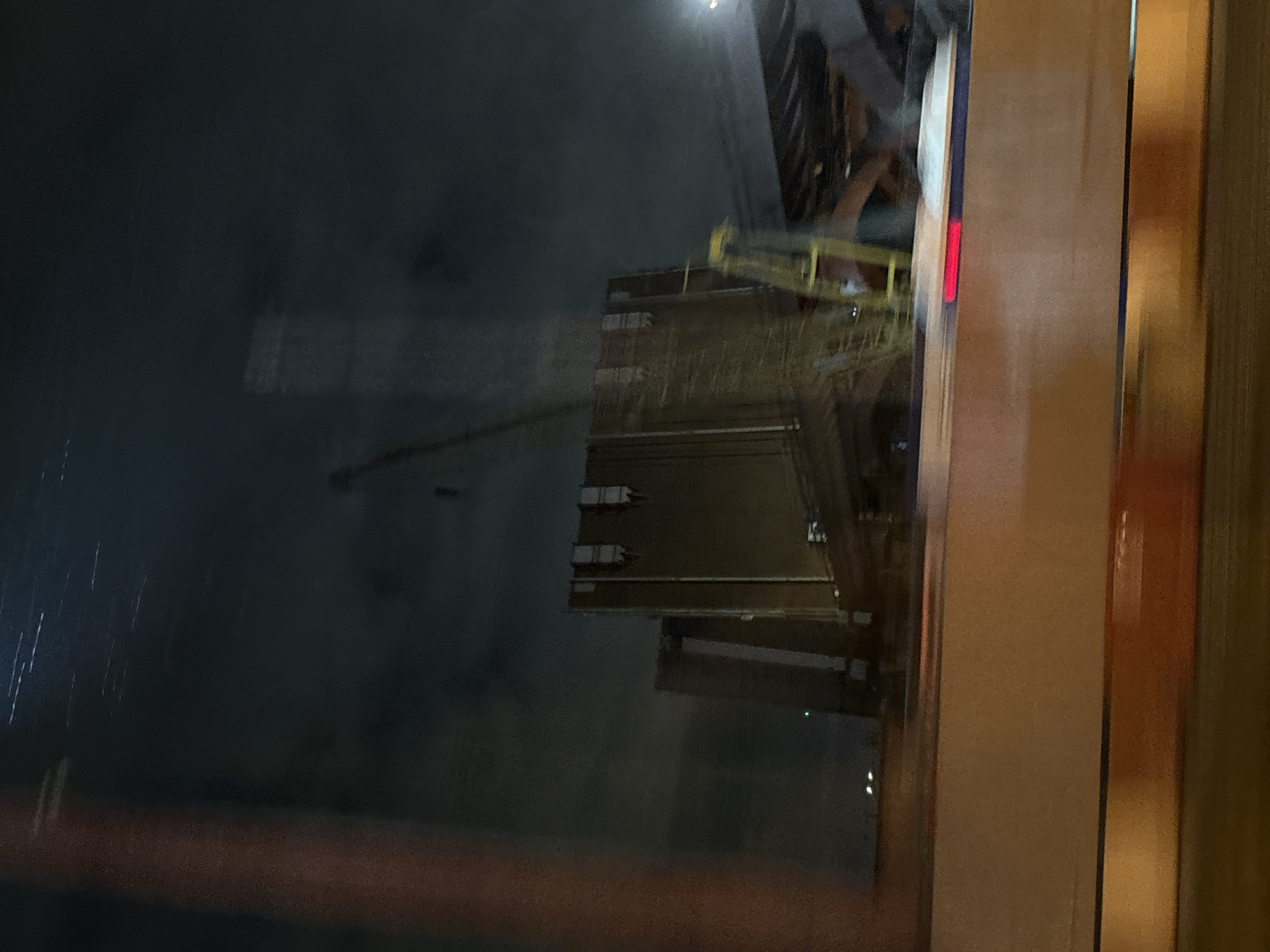 The image is a blurry nighttime photograph taken from a vehicle moving through Saint Petersburg, Russia. The primary subject is a large, multi-story industrial structure, possibly a grain elevator or silo complex, under construction or renovation. The structure is tall and rectangular, with a beige exterior and visible vertical supports and window-like openings. A large yellow construction crane is positioned to the left and slightly behind the main structure, with its arm extending upwards. There are also yellow scaffolding elements visible on the side of the building. The scene is illuminated by artificial lights, creating a dark and somewhat dramatic atmosphere. In the foreground, a beige barrier with a red light strip is visible, suggesting the photograph was taken from a roadway or bridge. The sky above is dark and appears to have some streaks of light, possibly from streetlights or other urban illumination reflecting on the clouds or the camera lens. The overall impression is one of a functional, industrial area at night, with signs of ongoing construction or development.