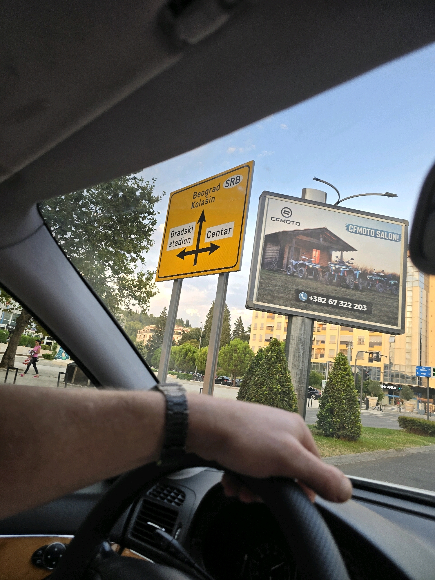 The image depicts a street scene in Podgorica, Montenegro, viewed from the interior of a vehicle. The driver's hand is visible on the steering wheel. In the foreground, a yellow directional sign points towards "Beograd SRB Kolasin," "Gradski stadion" (City Stadium), and "Centar" (Center). To the right of the directional sign, a billboard advertises "CFMOTO SALON!" with a phone number "+382 67 322 203." The billboard features an image of ATVs. In the background, trees, buildings, and a street with traffic lights are visible. A woman in a pink shirt and black pants is walking on the sidewalk to the left. The sky is a clear blue.