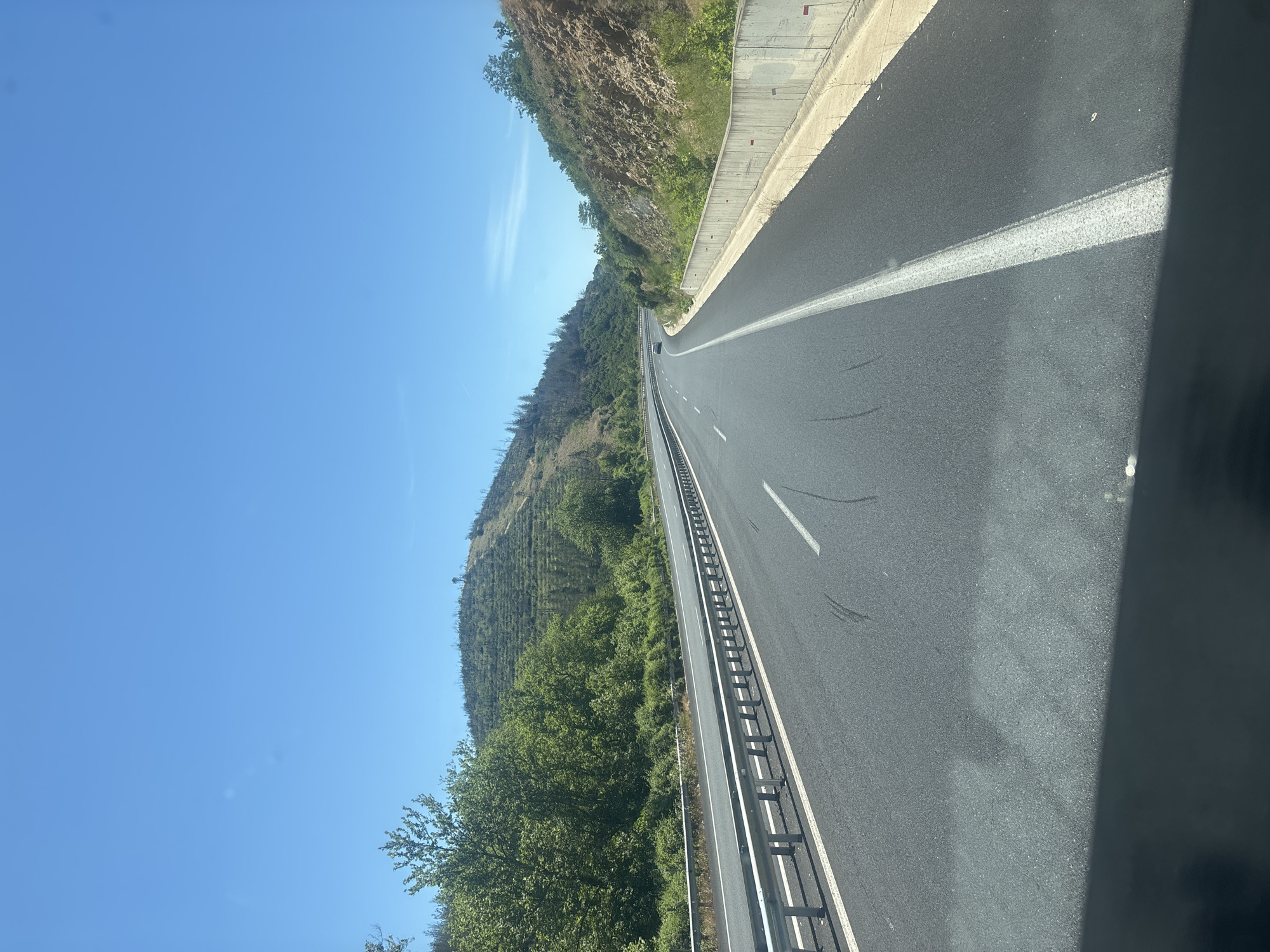 The image depicts a view from a vehicle traveling on a highway near Avra, Greece. The highway has multiple lanes separated by white lines. On the left side of the road, there is a metal guardrail and a steep, tree-covered hillside. The right side of the highway features a concrete barrier and a rocky embankment with sparse vegetation. The sky is clear and blue, with a few wispy clouds visible. The terrain appears hilly and somewhat arid, with evidence of past forest fires on some of the slopes, indicated by bare trees and scorched earth. One car is visible in the distance on the highway. The time of day appears to be daytime, likely mid-morning or early afternoon, given the bright sunlight and clear sky.