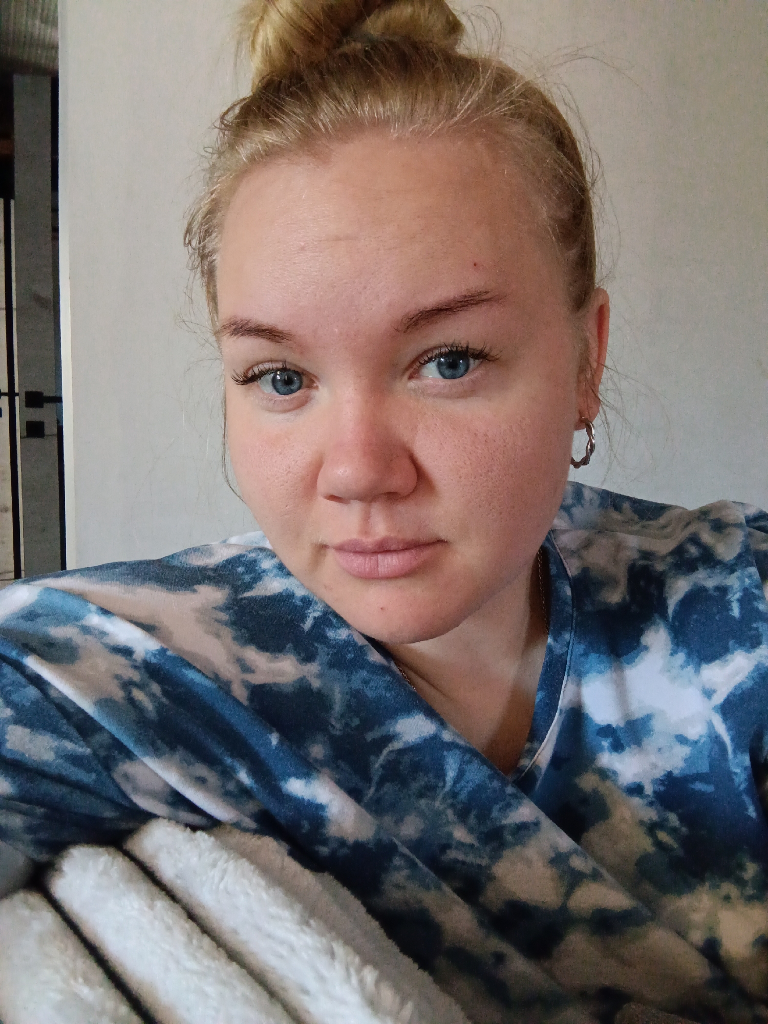 A medium close-up captures a young woman with fair skin and blue eyes. Her blonde hair is styled in a bun on top of her head. She is wearing a blue and white tie-dye shirt and a silver earring is visible on her left ear. She leans slightly to the left, resting on what appears to be a soft, white blanket or cushion. The background is a plain, light-colored wall with a glimpse of a doorframe to the left. The location is not discernible from the image and therefore cannot be specified as Фоки, Russia.