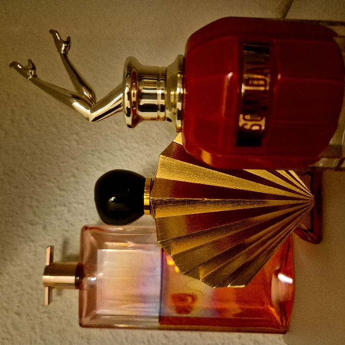 Three perfume bottles sit on a beige surface. The topmost bottle, "So Scandal!", is red and gold with a pair of gold legs sticking upwards. Below that is a gold, fan-shaped bottle with a black cap. The bottom bottle is rectangular and pink, with a gold cap resembling a small cross. The lighting is warm, causing the gold accents to shimmer.
