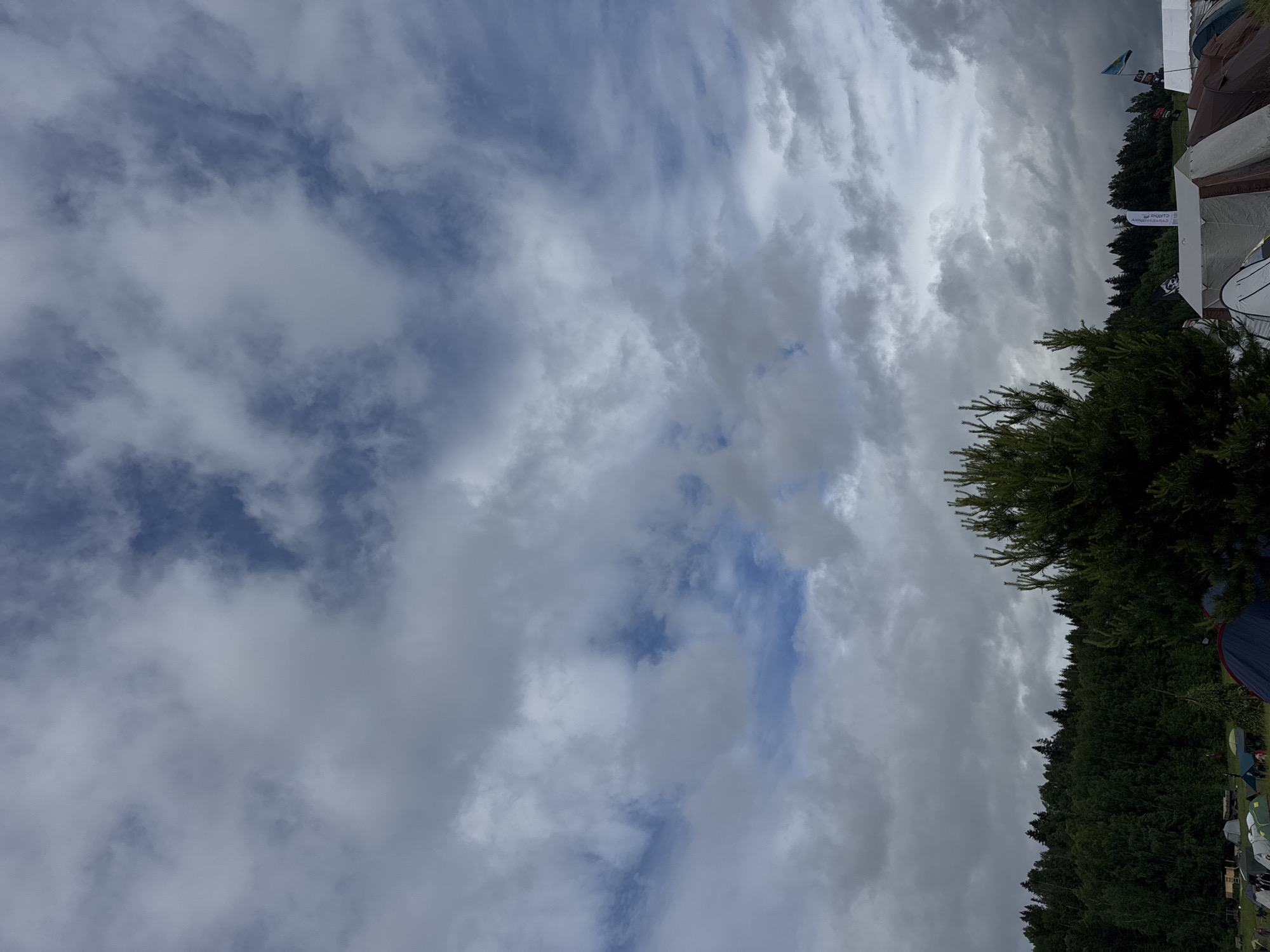 The image depicts an outdoor scene, likely an event or gathering, set under a dynamic cloudy sky. A dense line of coniferous trees forms the horizon, suggesting a natural or rural environment in Большая Кивара, Russia. Several tents are visible along the treeline on the right side of the frame, varying in size and color, some appearing to be camping tents, others possibly market stalls.
Prominently displayed among the tents is a white banner with dark text, which reads "ЯРМАРКА СУВЕНИРОВ" (Souvenir Market) in Cyrillic, clearly indicating the nature of the event. Above the tents, a blue and yellow flag, resembling the flag of Ukraine, is also partially visible.
The sky is a dominant feature, filled with a mix of thick grey and white clouds, with occasional glimpses of clear blue sky peeking through. The lighting suggests it is daytime, possibly overcast, contributing to a soft, diffuse illumination. No people are clearly identifiable in the image.
