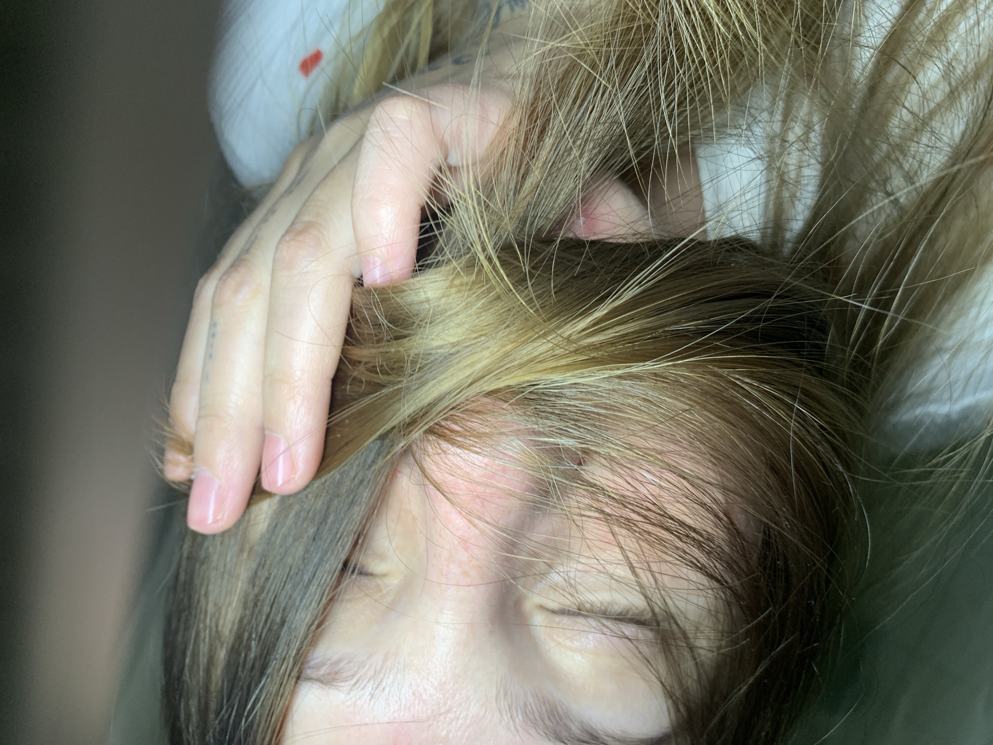 A close-up shot shows a person with their eyes closed. A hand with light skin and a tattoo on one of the fingers is partially visible, holding strands of light brown and blonde hair. The person's face is fair with freckles across the nose. The background is blurred and out of focus. There is no information available that indicates this image was taken in Chaikovsky, Russia.