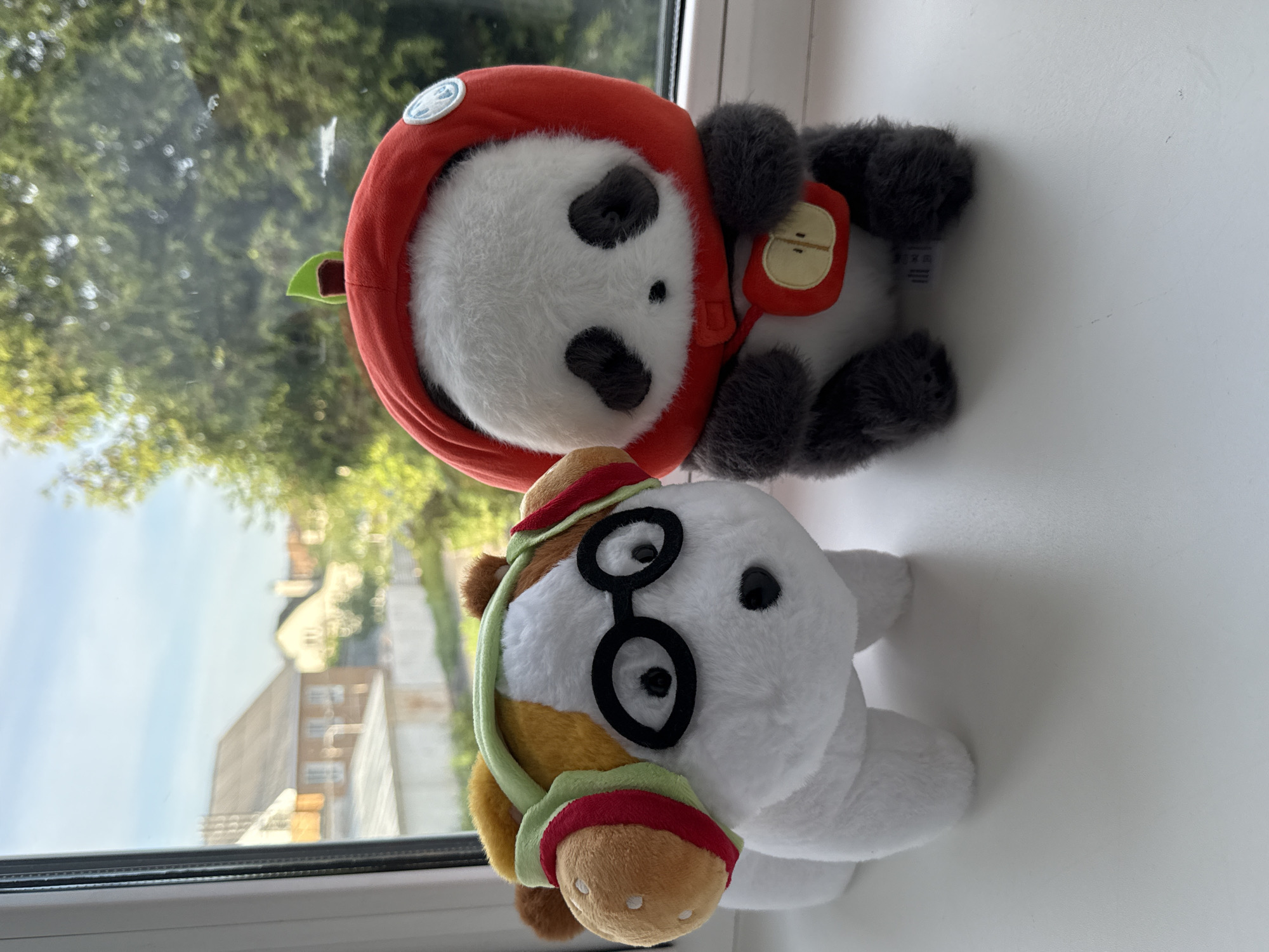Two plush toys sit on a white windowsill. The toy in the foreground is a white horse wearing black-rimmed glasses and a brown and red-trimmed hat and halter. Behind the horse is a panda bear plush wearing a red hood with a green apple stem detail and a red apple-shaped backpack. The background, visible through the window, features blurry foliage and buildings, suggesting an outdoor scene in Severnyi, Russia.