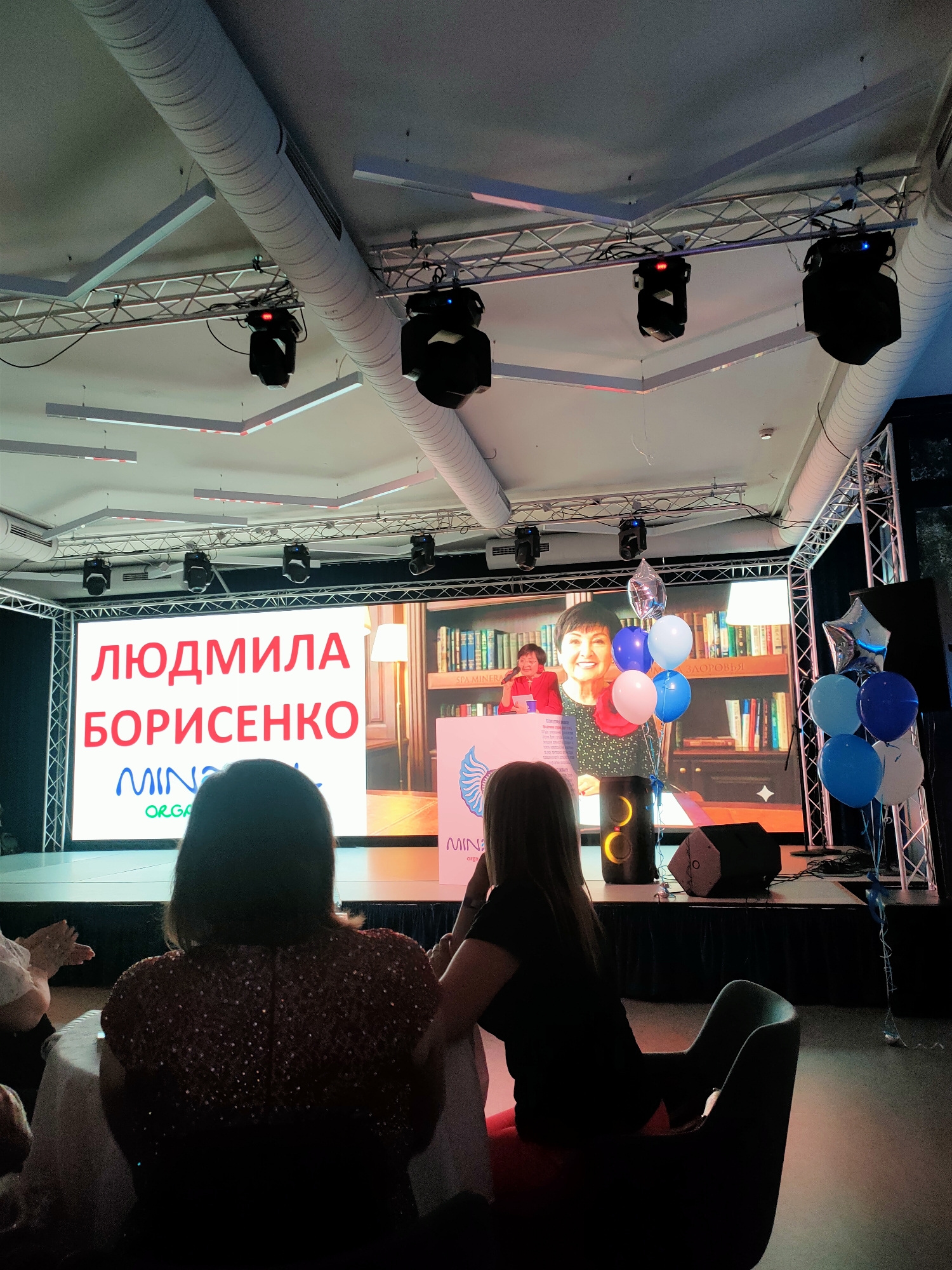 This image depicts an indoor event, likely a presentation or awards ceremony, with a large screen displaying text and an image. The screen prominently features the Cyrillic text "ЛЮДМИЛА БОРИСЕНКО" in large red letters, with smaller logos and text below. To the right of this text, a woman is visible on the screen, speaking at a podium. The background of the screen shows bookshelves. In the foreground, the backs of several attendees are visible, seated at tables and in chairs, facing the screen. Balloons, in shades of blue and white, are arranged on the stage area. The ceiling shows exposed ductwork and stage lighting equipment. The overall impression is of a formal or semi-formal gathering. The image is taken from a slightly elevated, rearward perspective, looking towards the stage. The lighting appears to be a mix of stage lights and ambient room light. There is no specific indication of the city of Kyiv, Ukraine in the image itself.