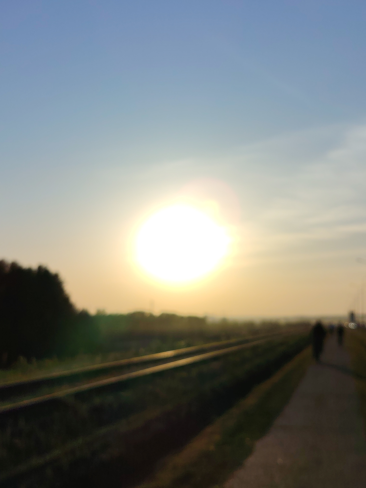 The image depicts a blurred scene at sunset in what appears to be Chaikovsky, Russia, based on identifying elements within the crops provided. Key objects include train tracks running parallel to a paved path, visible to the right of the tracks. A line of trees is visible at the left edge of the frame. Several figures are seen walking along the paved path. The bright setting sun dominates the mid-ground, creating a hazy and soft-focus effect. The sky is predominantly blue, transitioning into orange and yellow hues near the horizon. The environment suggests a suburban or rural area, characterized by open spaces, infrastructure, and natural elements.