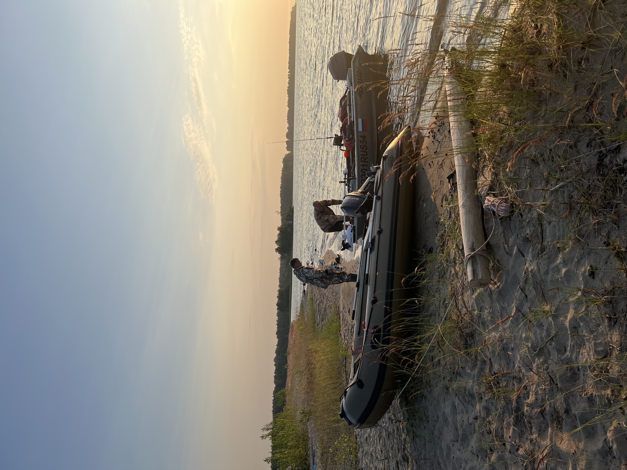 This image depicts a serene lakeside scene at sunset, likely in or around the city of Borovoe, Russia. Two inflatable boats are pulled ashore on a sandy beach, with tall grasses and driftwood scattered on the sand. In the background, a dense treeline borders the far bank of the calm body of water, and the sky is a gradient of pale blue and soft orange, indicating the late hour.
Two individuals, dressed in camouflage attire, are attending to the boats. One person is standing by the rear of the boat closer to the water, appearing to be engaged with something on the boat's stern. The second person is bent over near the front of the same boat, also seemingly occupied with an object or task. A fishing rod is visible in the rear boat, suggesting recreational activity. The water is gently lapping the shore, and the overall atmosphere is peaceful and quiet.