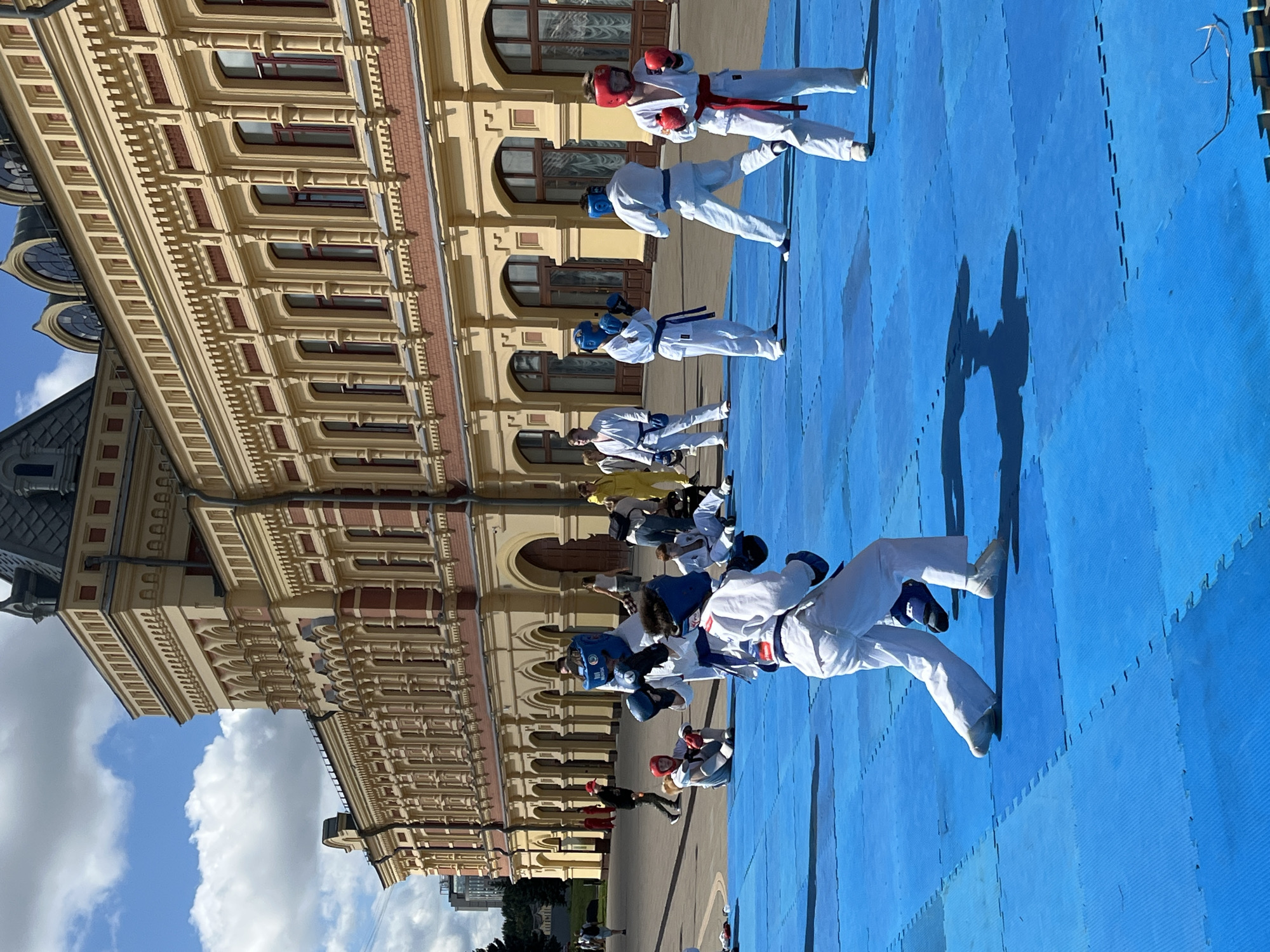 The image shows an outdoor training session for martial arts, likely karate or taekwondo, taking place on a blue mat in front of a large, ornate building. The building appears to be a historic structure, possibly a museum or administrative building, with a yellow facade, red brick accents, and arched windows. The sky is blue with scattered white clouds.
Several individuals are engaged in training activities. In the foreground, two individuals in white martial arts uniforms and protective gear (helmets and gloves) are facing each other, suggesting a sparring or practice match. The individual on the left is wearing a blue helmet and blue gloves, while the individual on the right is wearing a white uniform with a red belt and red gloves. Another individual in a white uniform with a blue belt and blue helmet is in the middle ground, also appearing to be practicing. To the right, another person in a white uniform with red gloves is standing ready. Further back, more people in similar attire are visible, some sitting on the mat and others standing. One person is wearing a yellow uniform. The scene is set in a city plaza or courtyard.
Based on the architectural style of the building, it is highly probable that this location is the Nizhny Novgorod State Art Museum (formerly the Rukavishnikov Estate) in Nizhny Novgorod, Russia. The distinctive yellow facade, the decorative elements above the windows, and the overall grandeur of the building are characteristic of this landmark. The presence of an outdoor event like a martial arts training session in such a prominent public space is also plausible for this area.